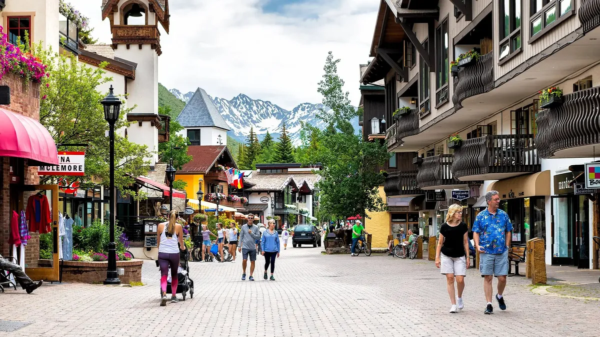 This image showcases a vibrant street scene lined with charming buildings, possibly in a mountain resort town. The architecture features elements like balconies, flower boxes, and varied rooflines, adding character to the properties. People are seen strolling and enjoying the outdoors, suggesting a lively and pedestrian-friendly environment.