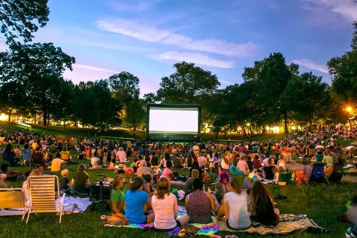 This image showcases a large outdoor movie screening in a park-like setting. A crowd sits on the lawn facing a large inflatable screen, suggesting a community event or amenity. The surrounding trees and twilight sky create a pleasant atmosphere, likely appreciated by residents and contributing to the area's appeal.