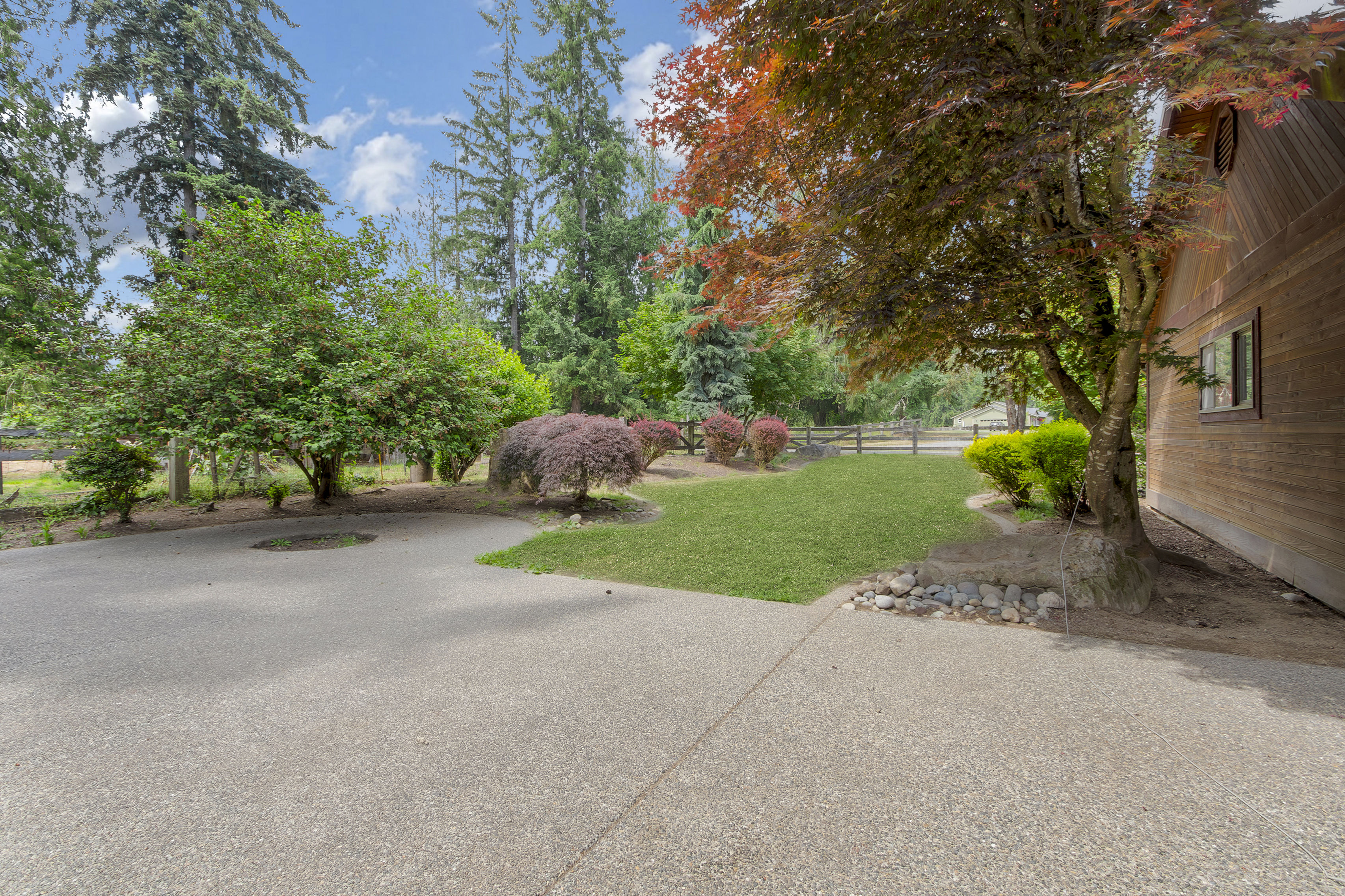 This image showcases a well-maintained yard with a mix of mature trees, shrubs, and a patch of green lawn. A concrete driveway leads up to the property, and a wooden fence can be seen in the background, suggesting a spacious and private setting. The overall impression is one of tranquility and natural beauty.