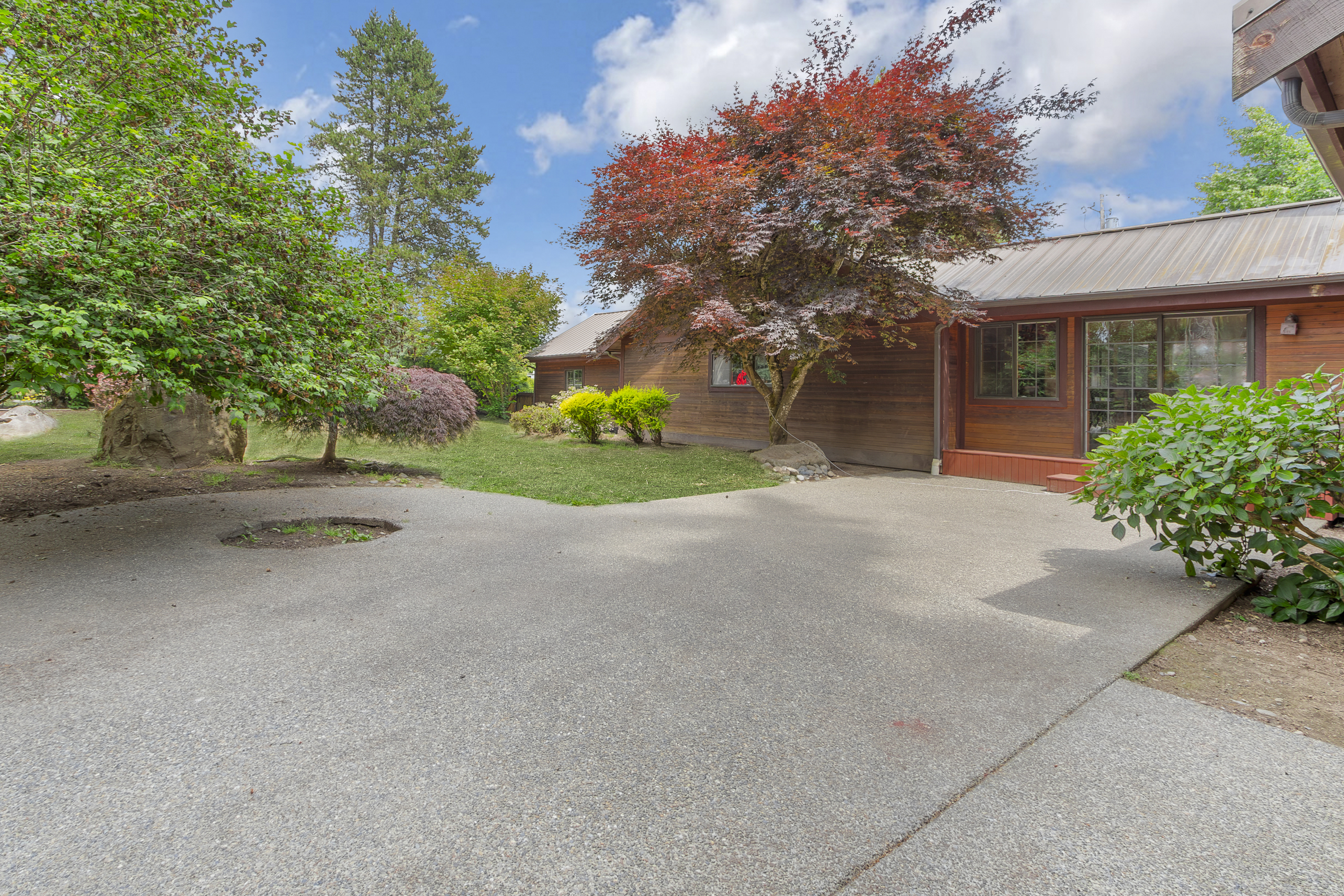This exterior shot showcases a well-maintained yard and garden area. A spacious concrete driveway leads to a wooden house with a metal roof, complemented by mature trees, including a vibrant red maple. The scene presents a serene and inviting outdoor space, perfect for relaxation and outdoor activities.