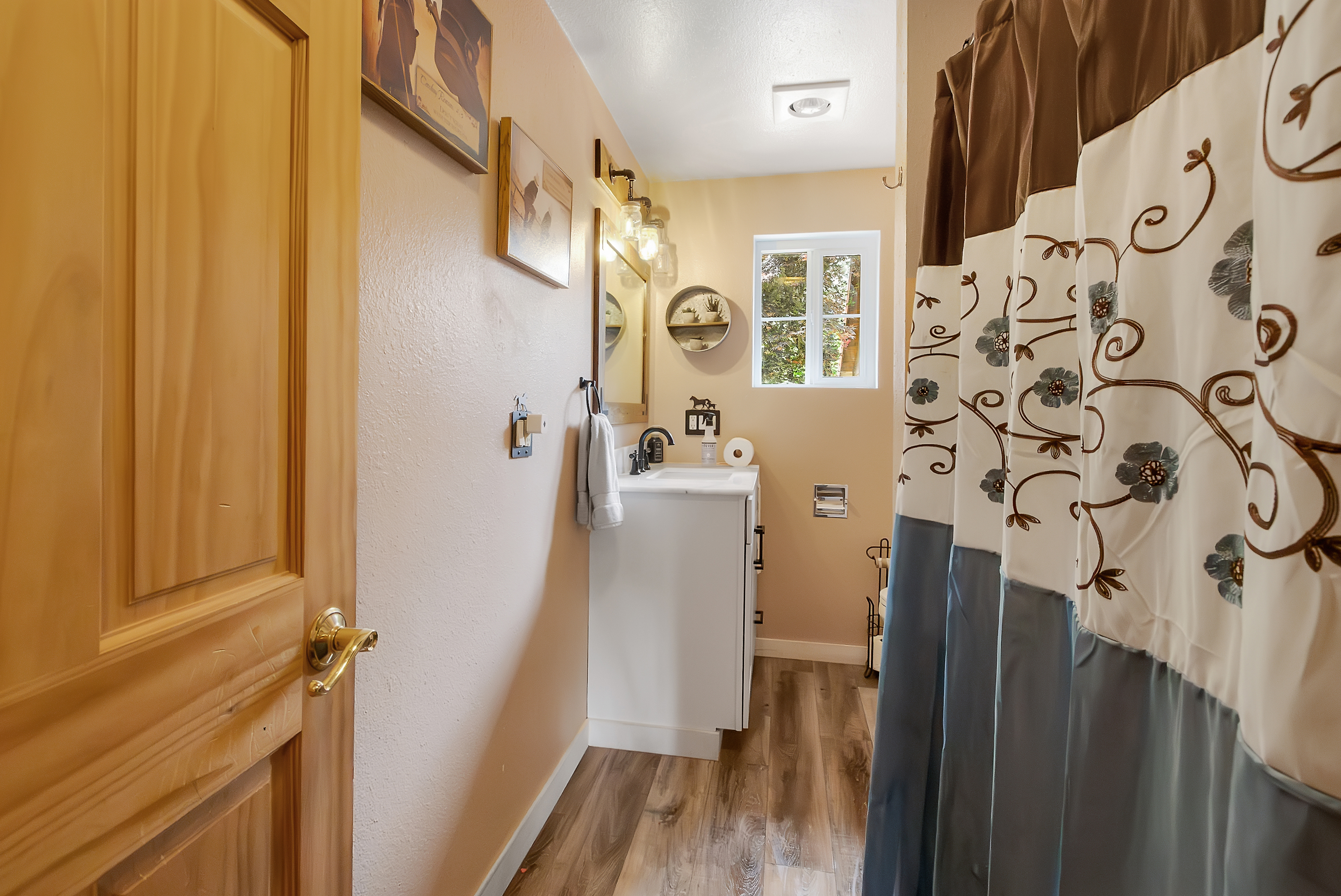 This is a cozy bathroom featuring a white vanity with a dark faucet, complemented by a framed mirror and decorative lighting. The walls are painted in a warm, neutral tone, and the flooring is wood-style laminate. A patterned shower curtain adds a touch of style, and a small window provides natural light.