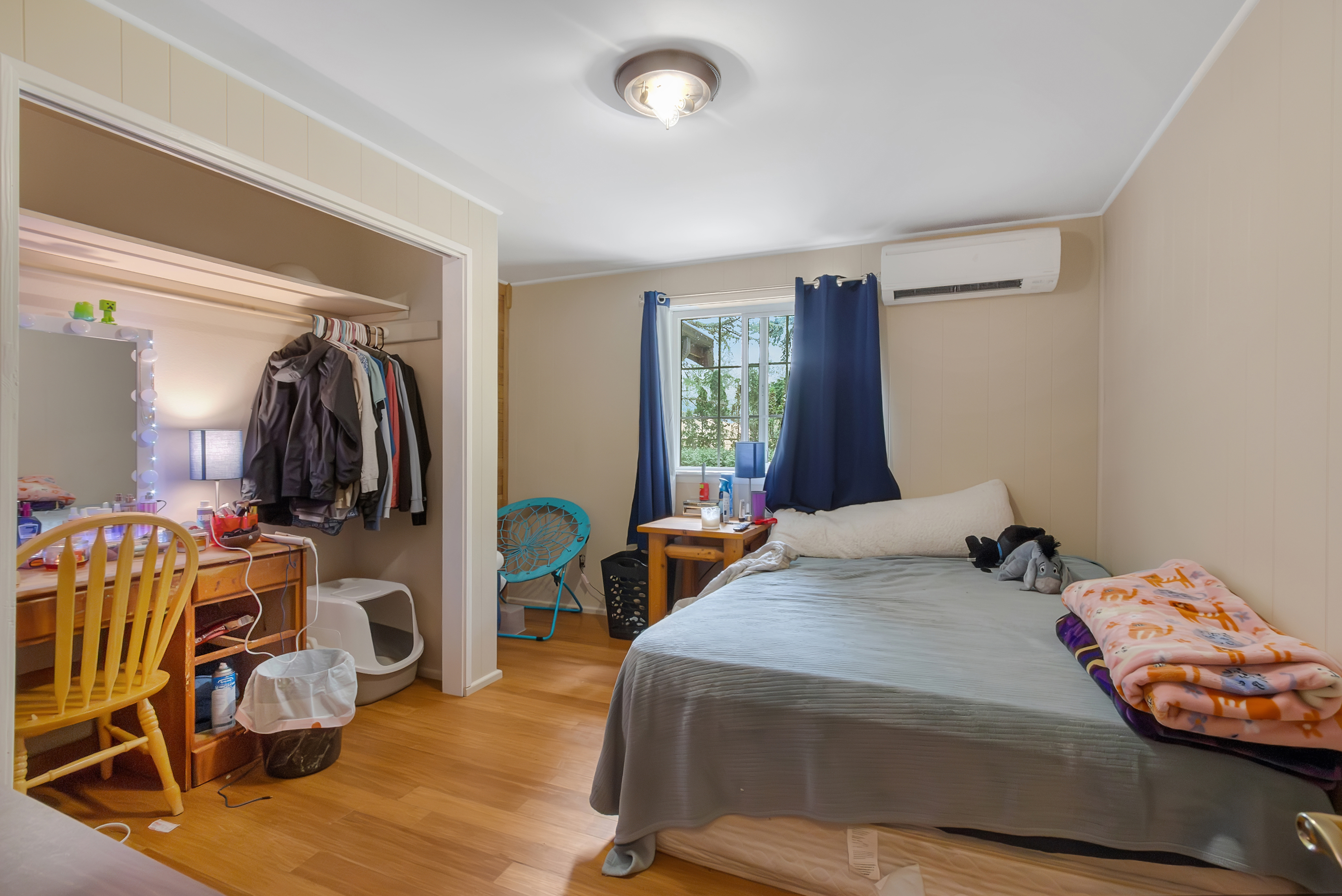This is a cozy bedroom featuring a bed with a light blue comforter and several blankets. The room has light-colored walls and wood flooring. A desk and chair are visible in the corner, along with a closet containing clothes. The room is lit by a ceiling light and a lamp on the desk, creating a warm and inviting atmosphere.