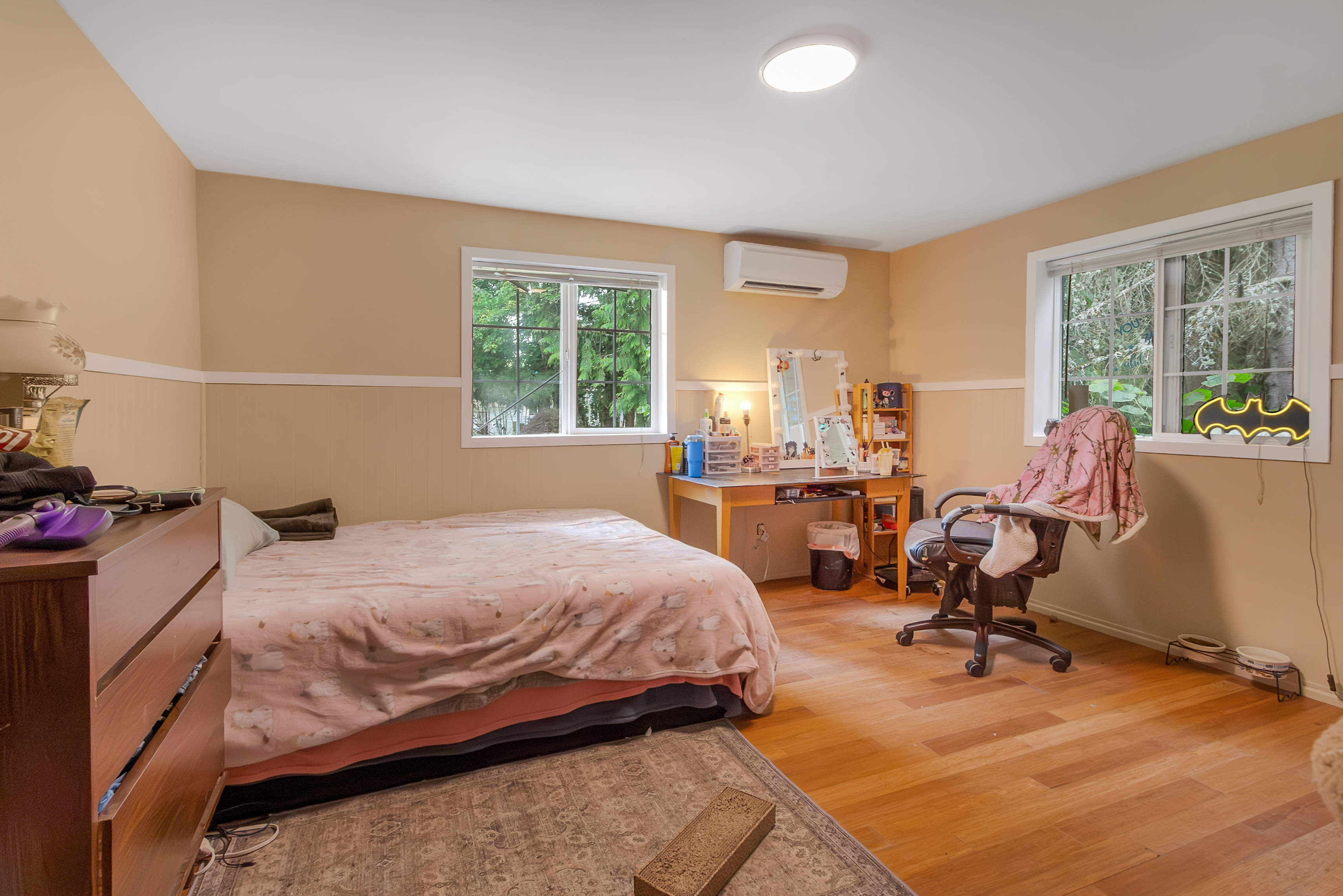 This is a cozy bedroom featuring a bed with a pink patterned comforter, a wooden dresser, and a desk area with a mirror and various items. The room has light beige walls with white trim, hardwood floors, and two windows that provide natural light. A Batman-shaped light fixture is visible near one of the windows, adding a unique touch to the space.