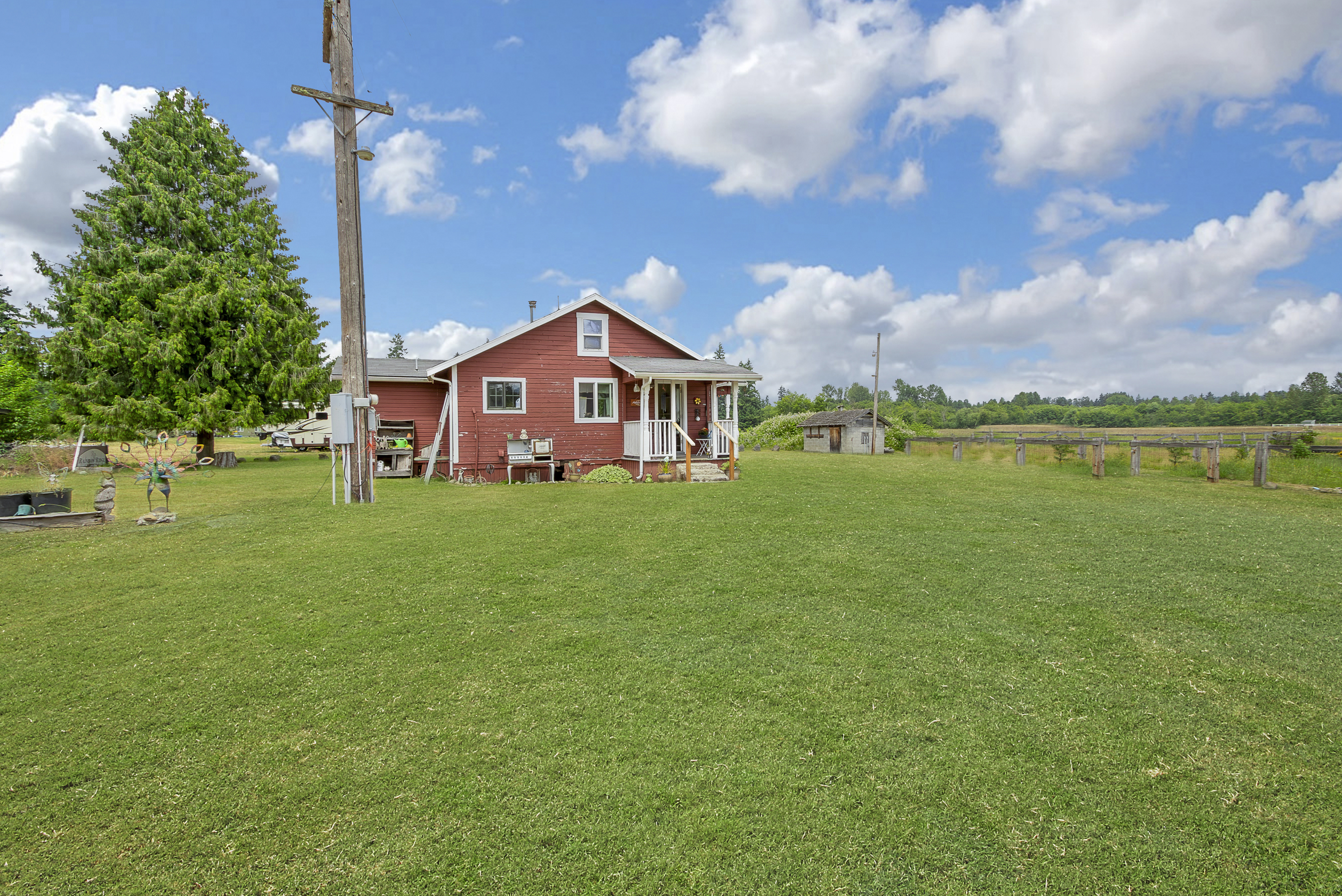 This image showcases the front exterior of a charming red farmhouse-style home with a welcoming porch. The house is surrounded by a large, well-maintained green lawn, and a wooden utility pole stands nearby. The sky is bright blue with fluffy white clouds, creating a picturesque and inviting scene.