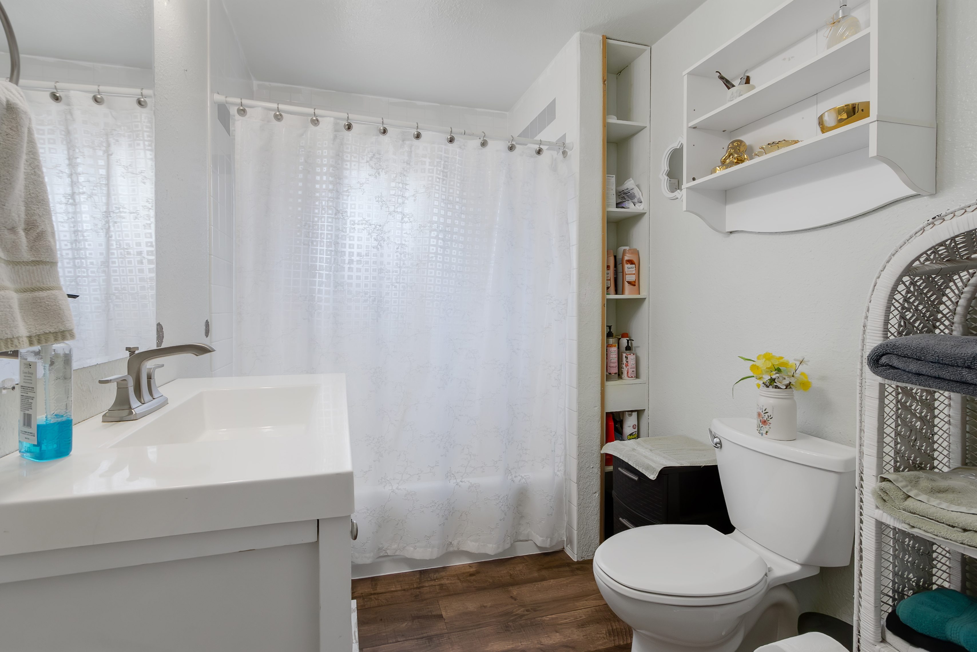 This is a well-lit bathroom featuring a white vanity with a modern faucet, a shower with a white curtain, and a toilet with a decorative vase on top. A built-in shelving unit and a white shelf provide storage space. The dark wood-look flooring adds a touch of warmth to the otherwise bright and clean space.