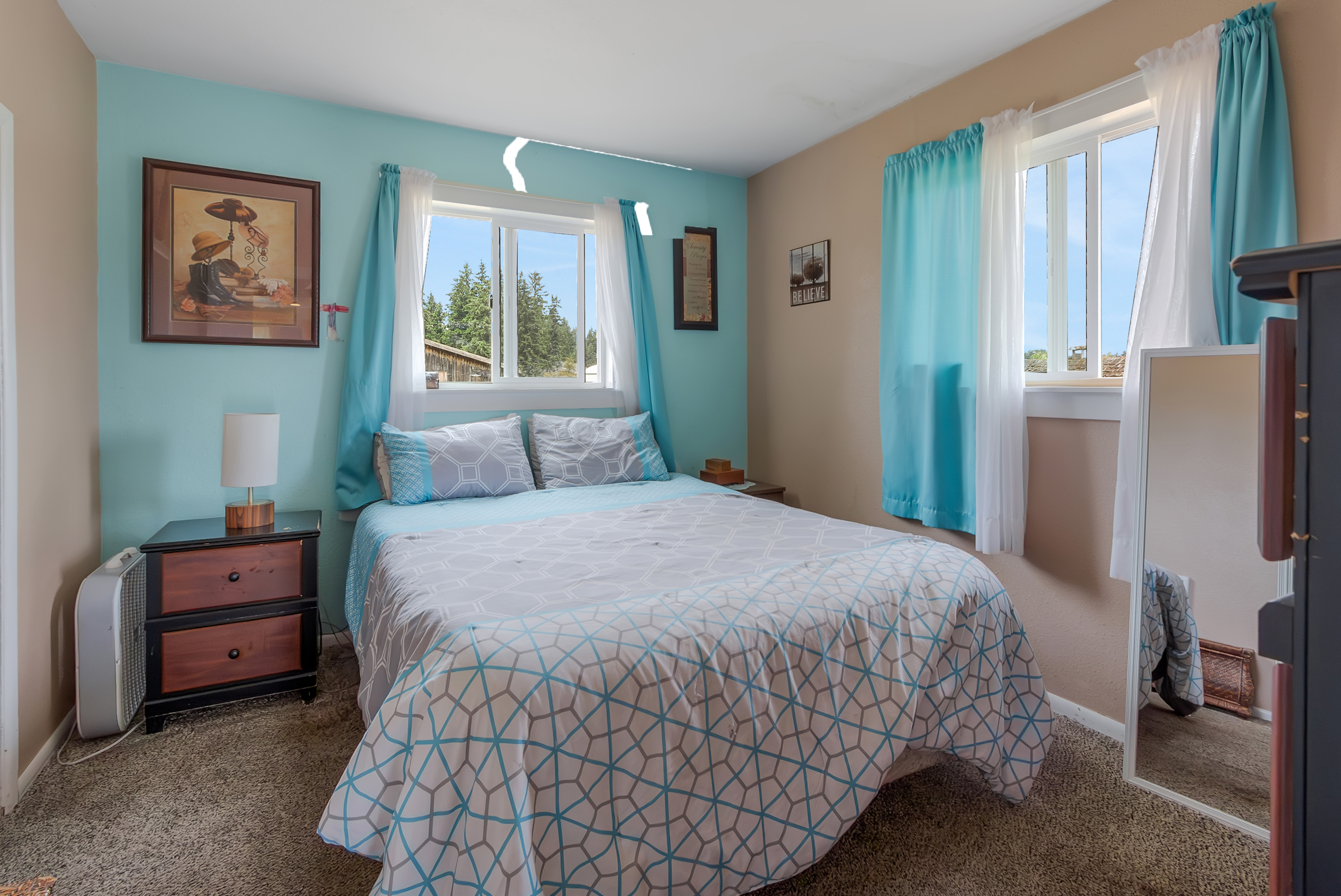 This is a cozy bedroom featuring a bed with a patterned comforter, two windows with light blue curtains, and a wooden nightstand with a lamp. The walls are painted in a combination of light blue and beige, creating a calming atmosphere. The room is carpeted and appears well-lit, offering a comfortable and inviting space.