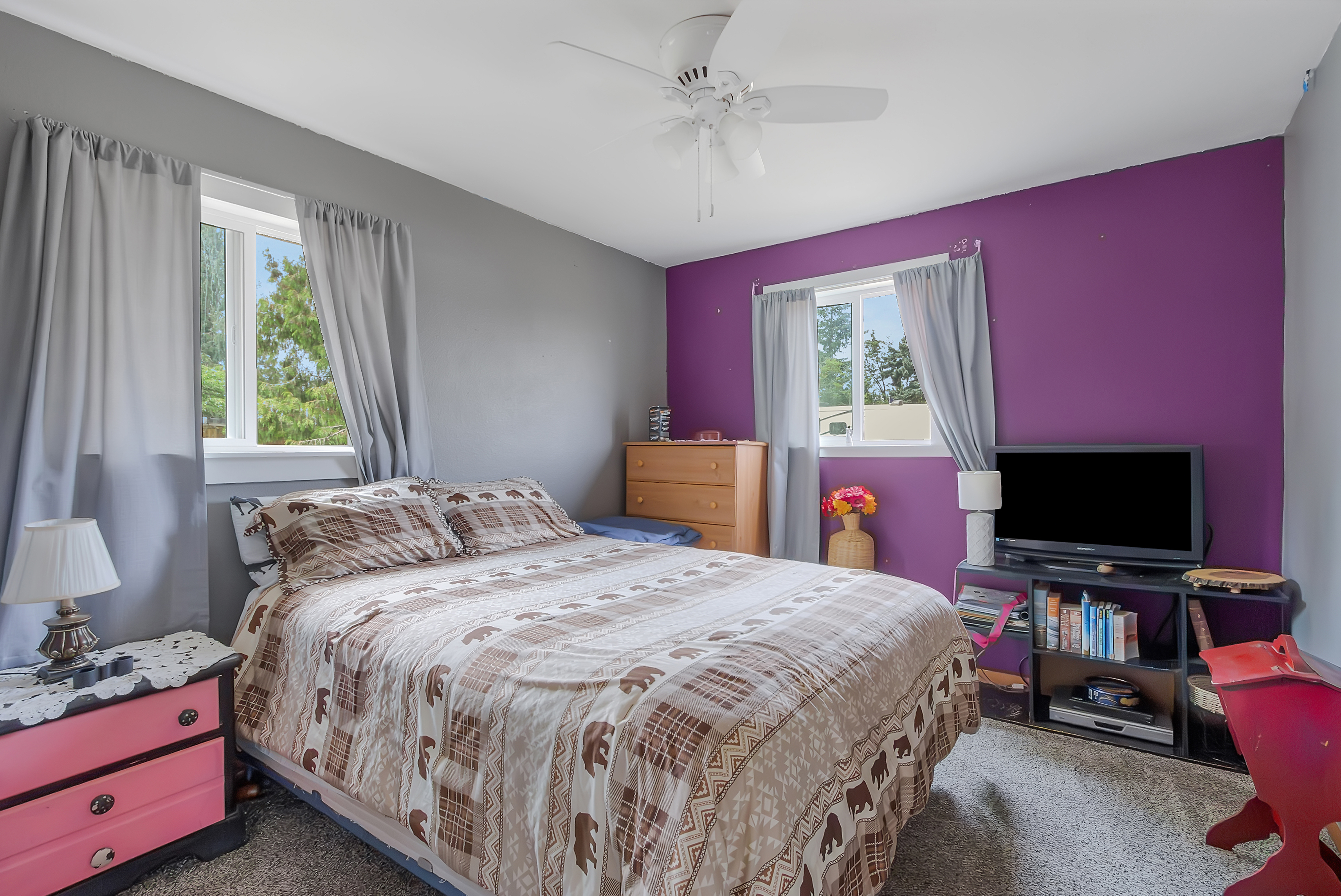 This is a cozy bedroom featuring a bed with a patterned comforter, a pink nightstand with a lamp, and a TV on a black stand. The walls are painted in a combination of gray and purple, and natural light enters through two windows with gray curtains. The room has a comfortable and inviting atmosphere.