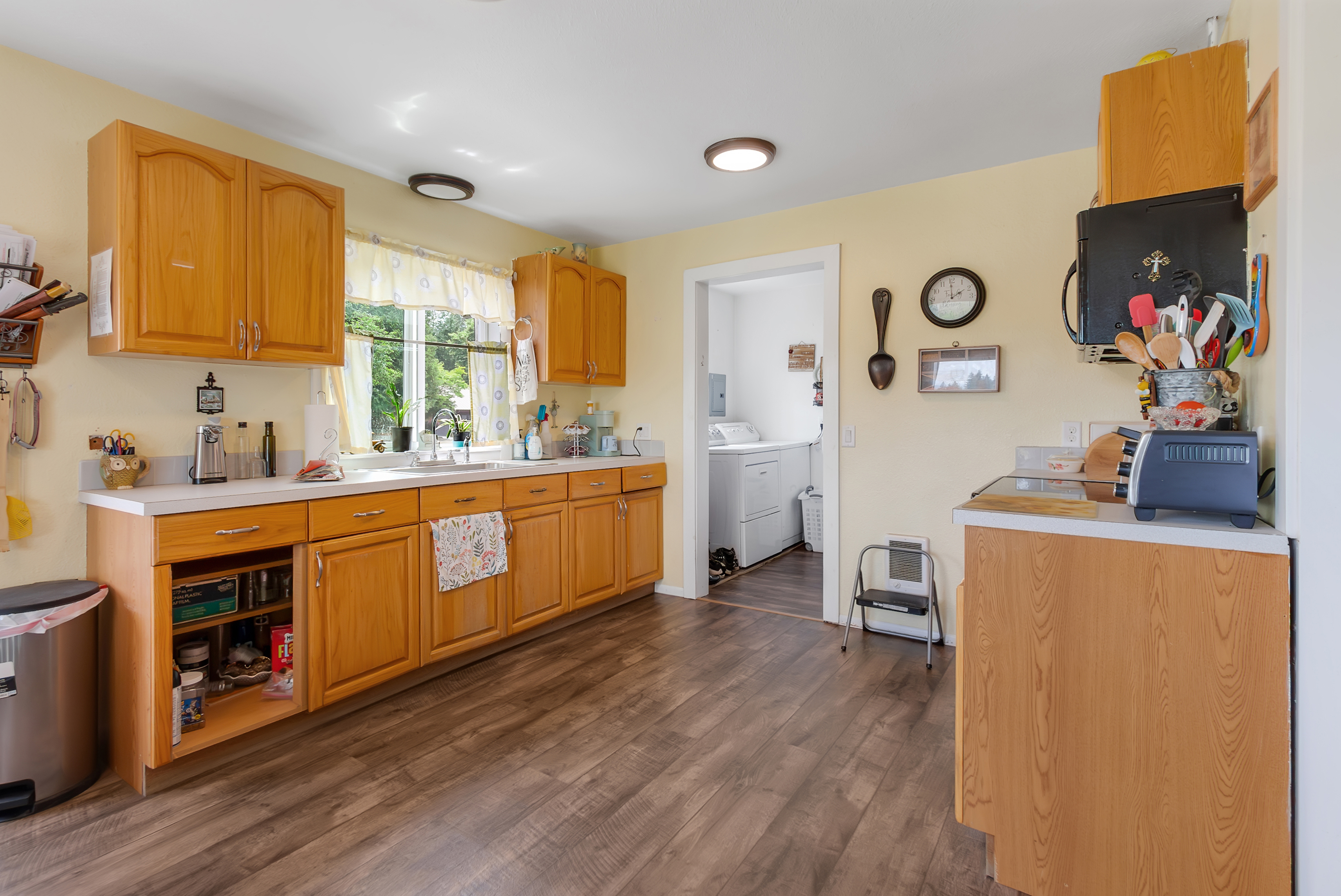 This is a warm and inviting kitchen featuring wood cabinetry, light yellow walls, and wood-look flooring. The kitchen is well-lit with recessed lighting and natural light from a window above the sink. Appliances include a toaster oven and a microwave, and there is an adjacent laundry room visible through a doorway, suggesting convenience and functionality.