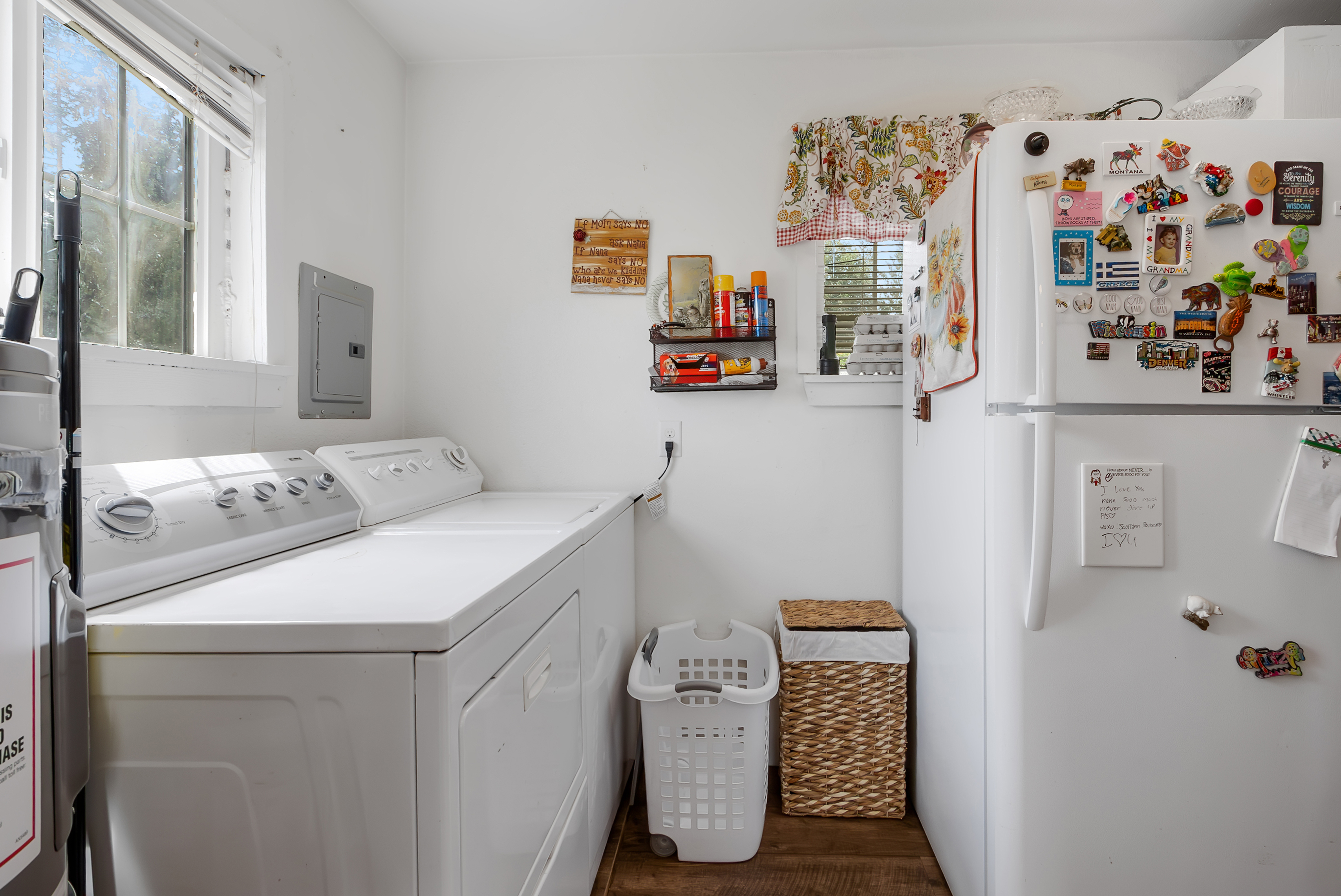 This is a well-lit laundry room featuring a white washer and dryer set, positioned beneath a window that allows natural light to fill the space. A white plastic laundry basket and a woven hamper provide storage solutions. A refrigerator adorned with magnets and notes stands to the right, adding a personal touch to the functional space.