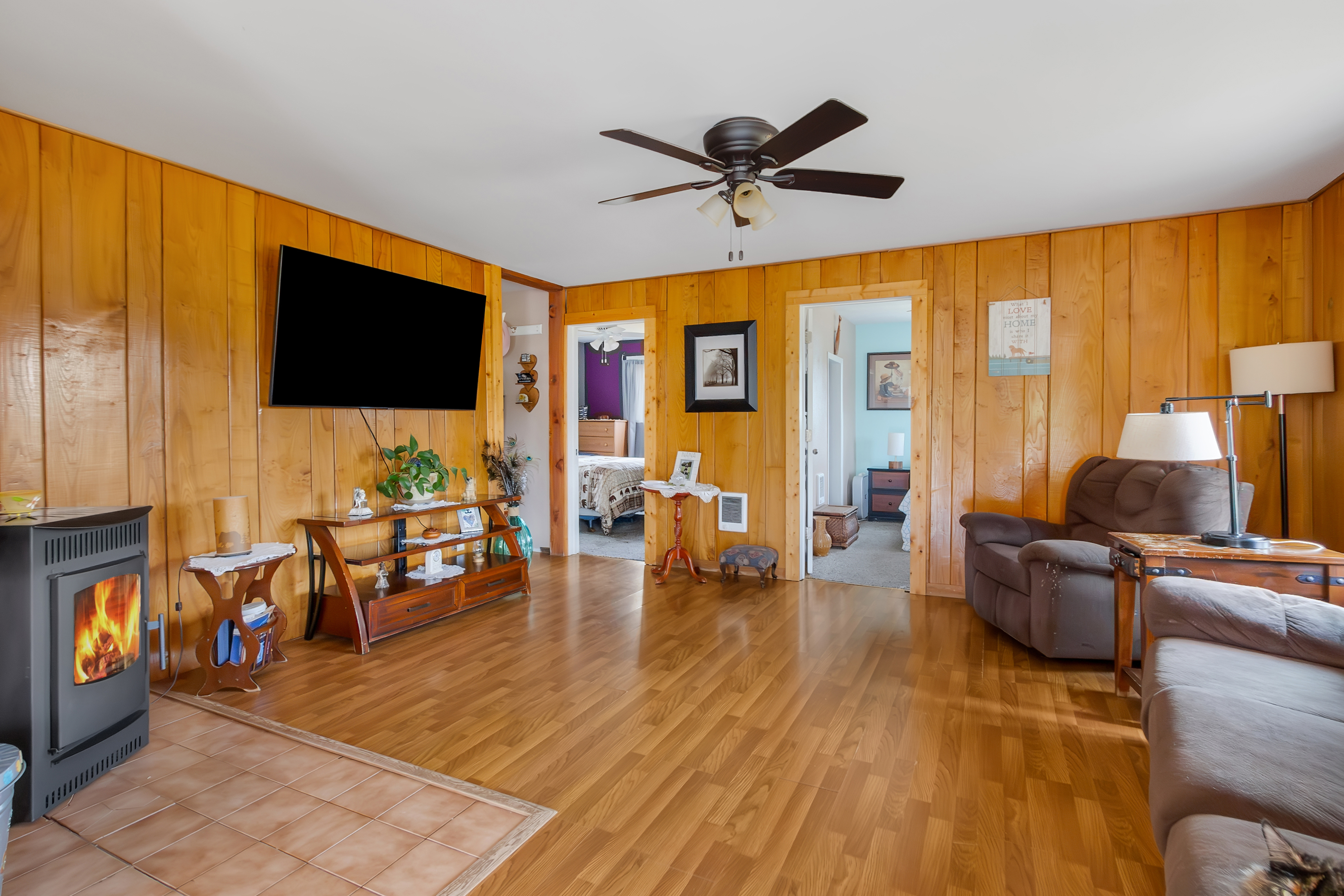 This is an interior shot of a cozy living room featuring wood-paneled walls and hardwood floors. A large flat-screen TV is mounted on the wall above a wooden entertainment center. The room includes comfortable seating, a wood-burning stove, and doorways leading to other rooms, creating a warm and inviting atmosphere.