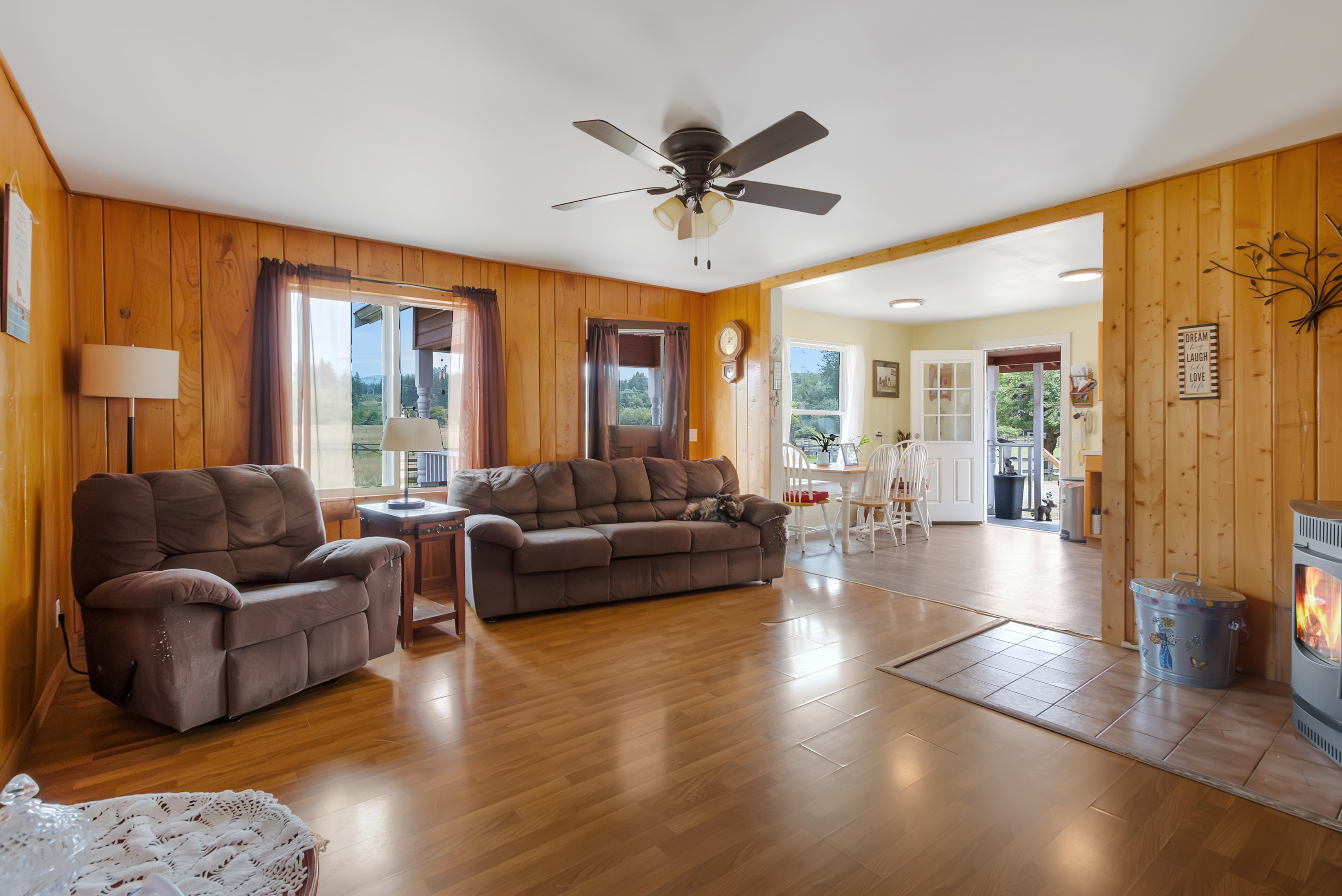 This inviting living room features warm wood paneling, a ceiling fan, and comfortable brown upholstered seating including a sofa and armchair. Natural light streams in through the windows, highlighting the hardwood floors. The space flows seamlessly into the adjacent dining area, creating an open and airy feel.