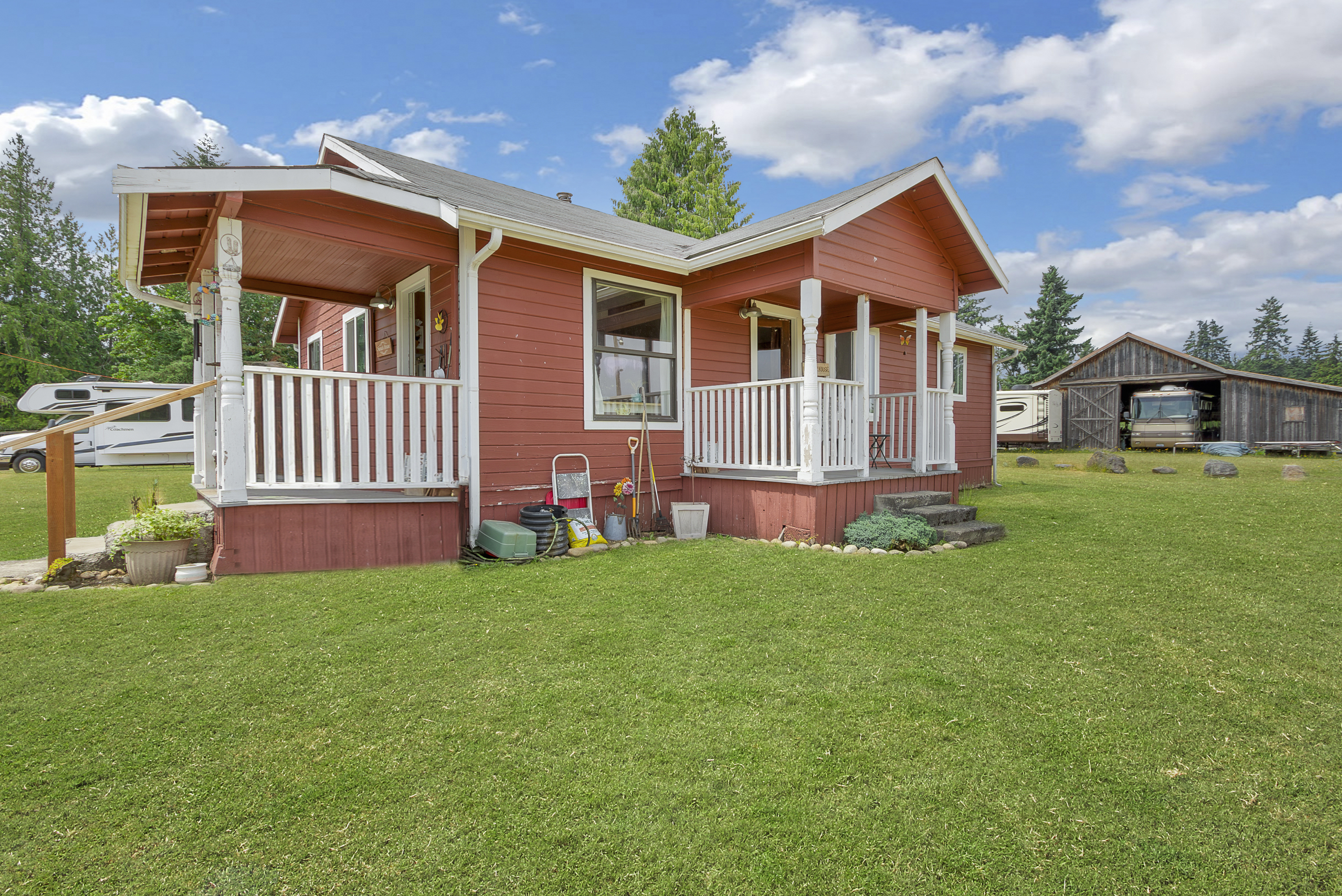 This is a front exterior view of a charming red house with white trim and a welcoming porch. The house is surrounded by a well-maintained green lawn, and there's a barn and RV visible in the background, suggesting ample outdoor space. The overall impression is one of a cozy and well-cared-for property.