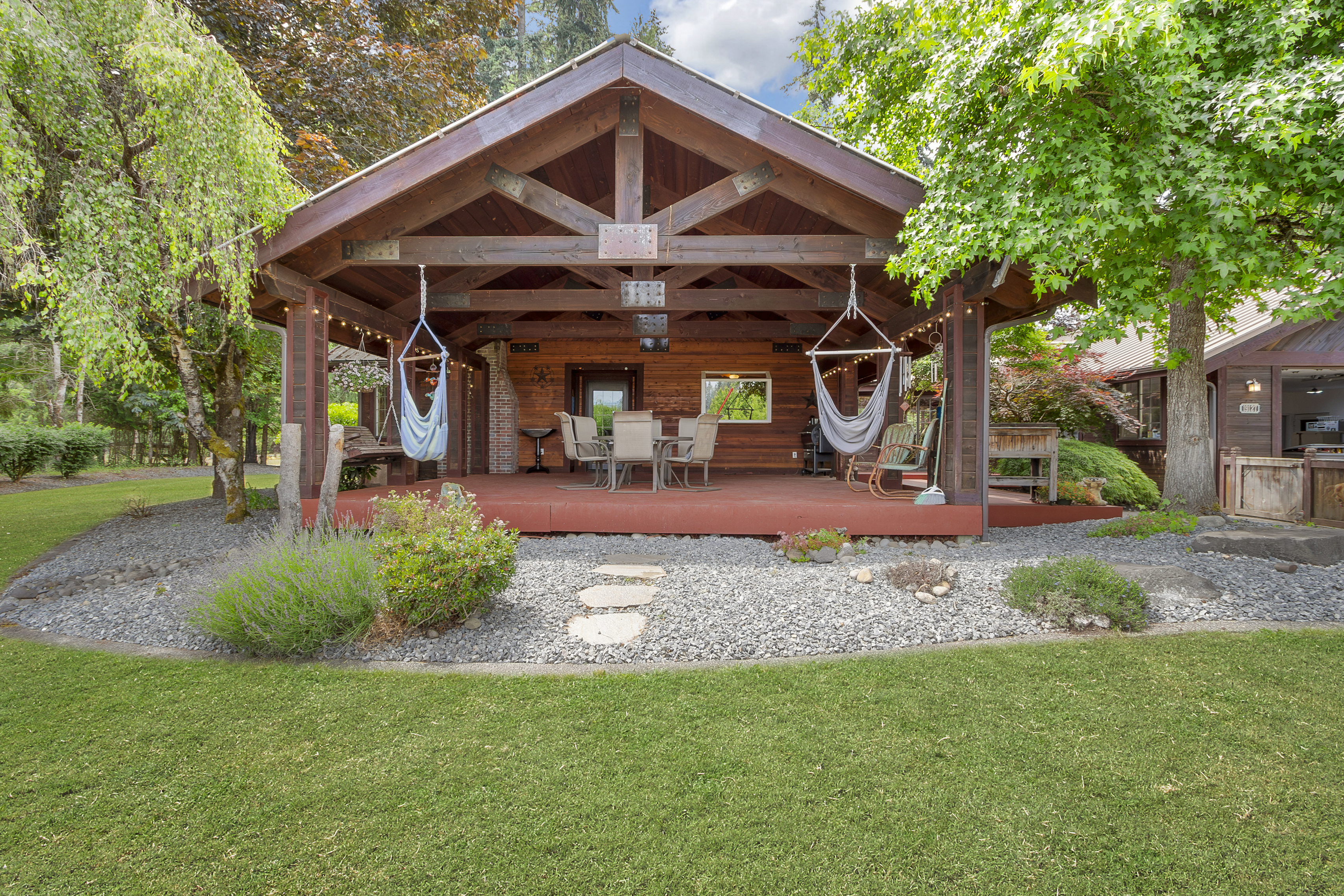 The image showcases a charming outdoor patio/deck area, featuring a rustic wooden structure with exposed beams and a cozy seating arrangement. Hammock chairs hang invitingly, and string lights add a warm ambiance. The surrounding landscape includes a well-maintained lawn, gravel pathways, and lush greenery, creating a serene and inviting outdoor living space.