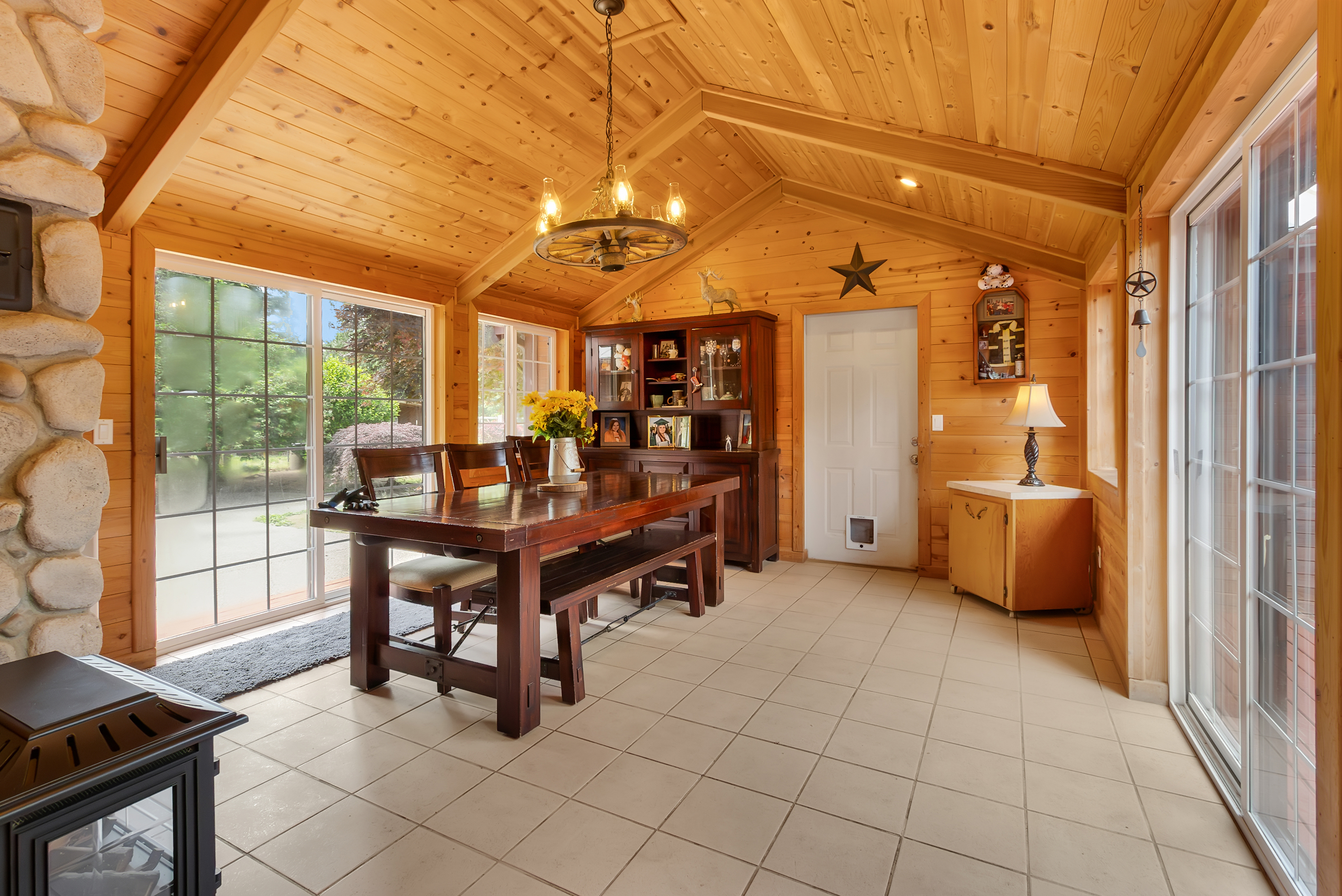 This is an interior shot of a dining room with a rustic and warm ambiance. The room features wood-paneled walls and ceiling, a large wooden dining table with benches and chairs, and a built-in hutch. Natural light floods the space through large windows and sliding glass doors, creating an inviting atmosphere.