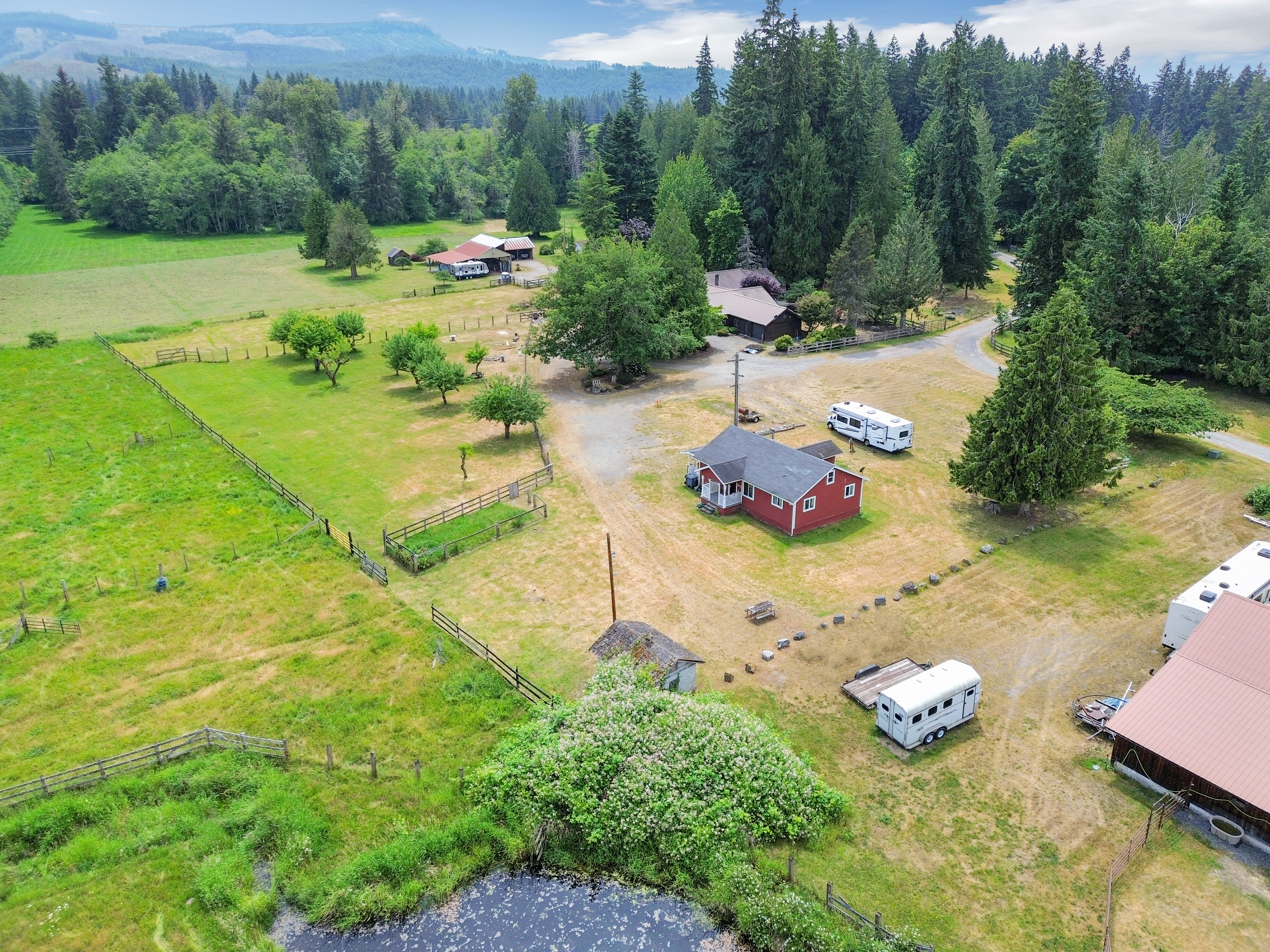 This aerial shot showcases a charming rural property featuring a red house with a dark roof, surrounded by lush greenery and mature trees. The property includes a well-maintained lawn, a small pond, several outbuildings, and fenced pastures, suggesting a farm or homestead. The overall impression is one of tranquility and spaciousness, ideal for a buyer seeking a peaceful country lifestyle.