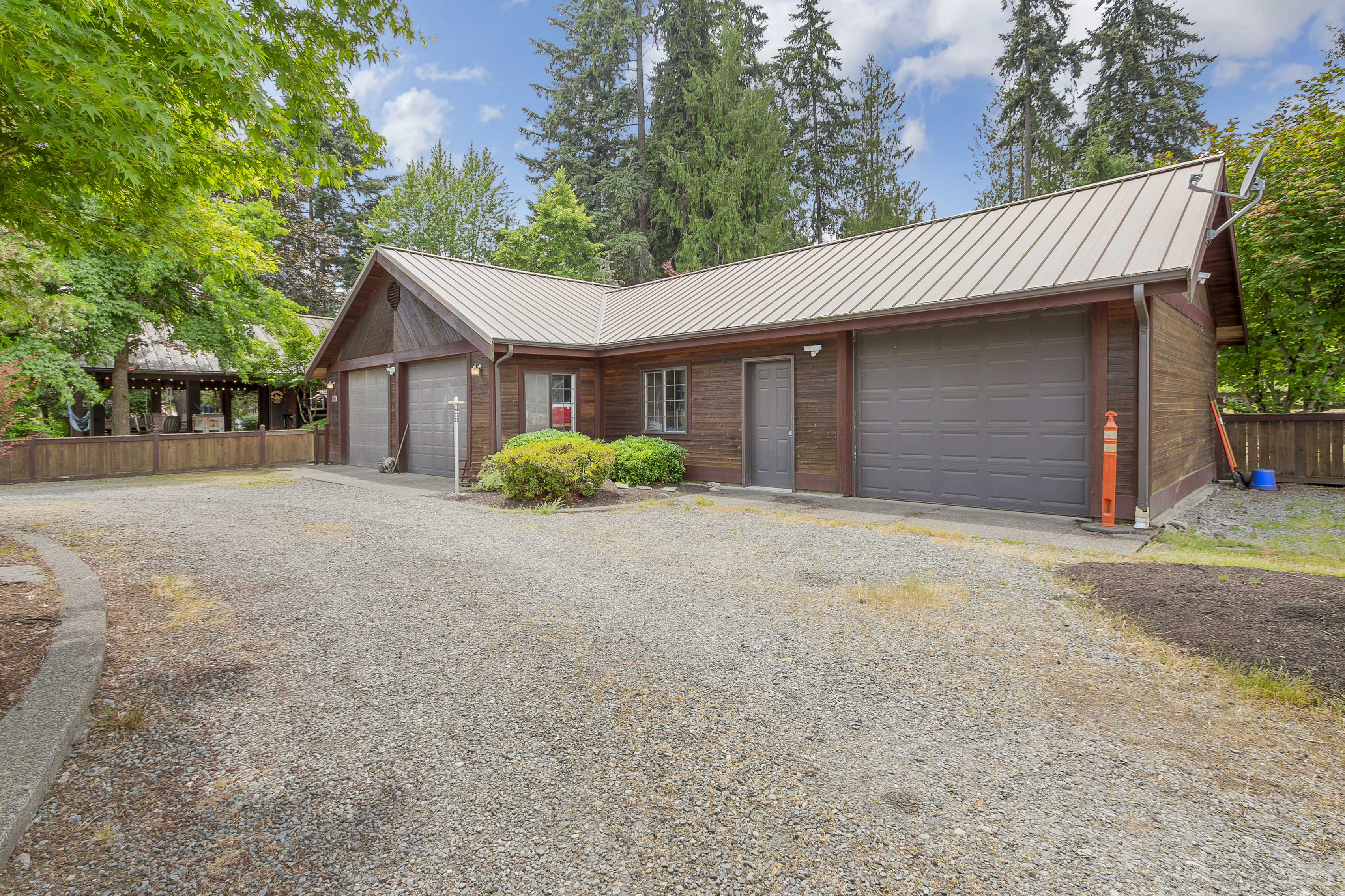 The image showcases a detached garage with a rustic, wood-paneled exterior and a metal roof. It features multiple garage doors, suggesting ample parking or storage space. The gravel driveway and surrounding greenery add to the property's charm, creating a functional yet aesthetically pleasing space.