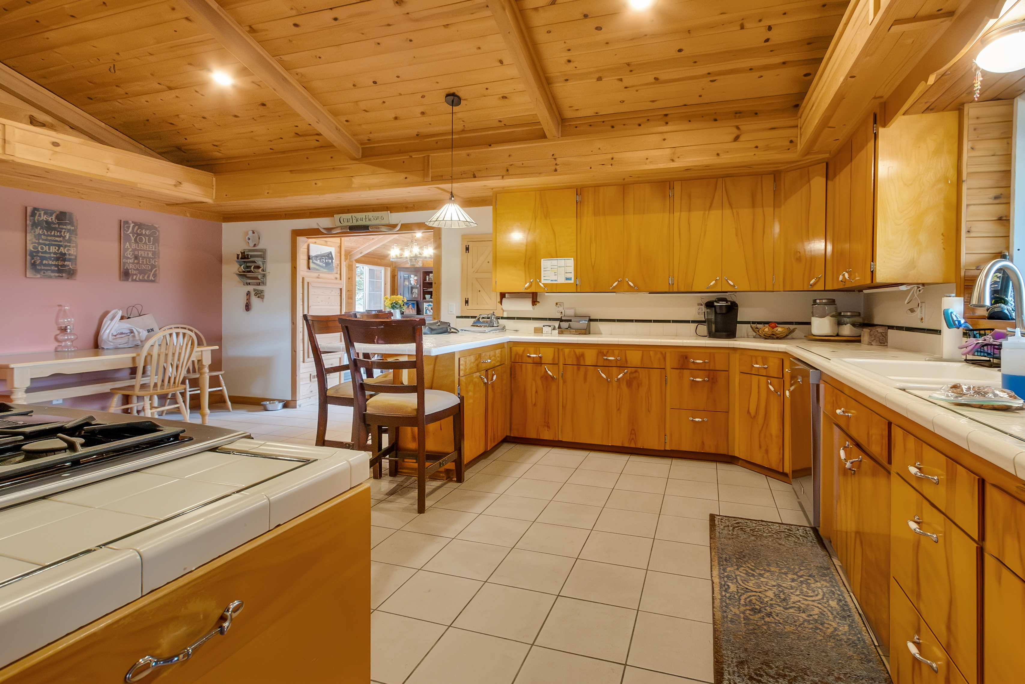 This is a warm and inviting kitchen with wood cabinetry and a matching wood plank ceiling. The kitchen features a white tile countertop and backsplash, with a vintage-style gas range visible in the foreground. The space is well-lit and appears to be part of a larger, open-concept living area.