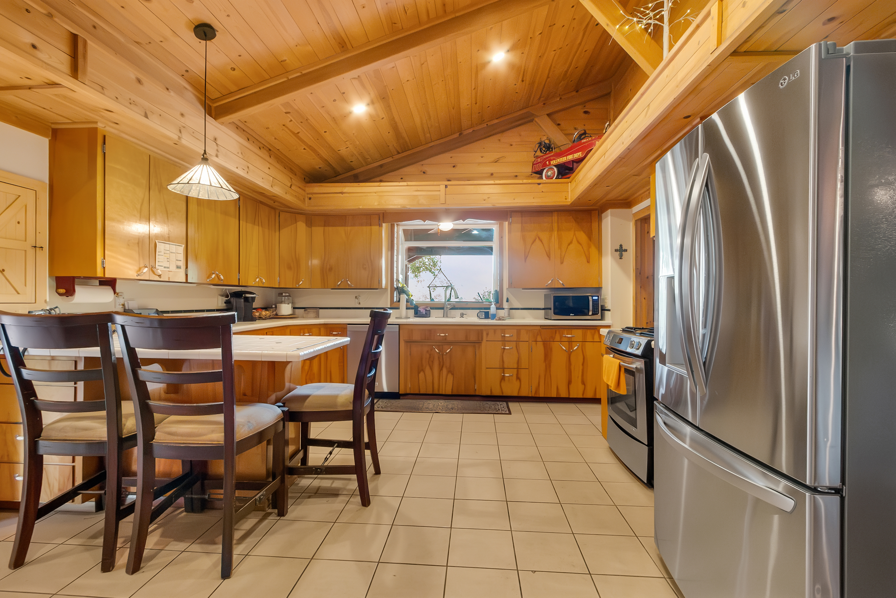 This is a warm and inviting kitchen with wood cabinetry and a matching wood-paneled ceiling. Stainless steel appliances, including a large refrigerator and oven, provide modern convenience. A central island with bar seating offers a casual dining space, and a window above the sink provides natural light.