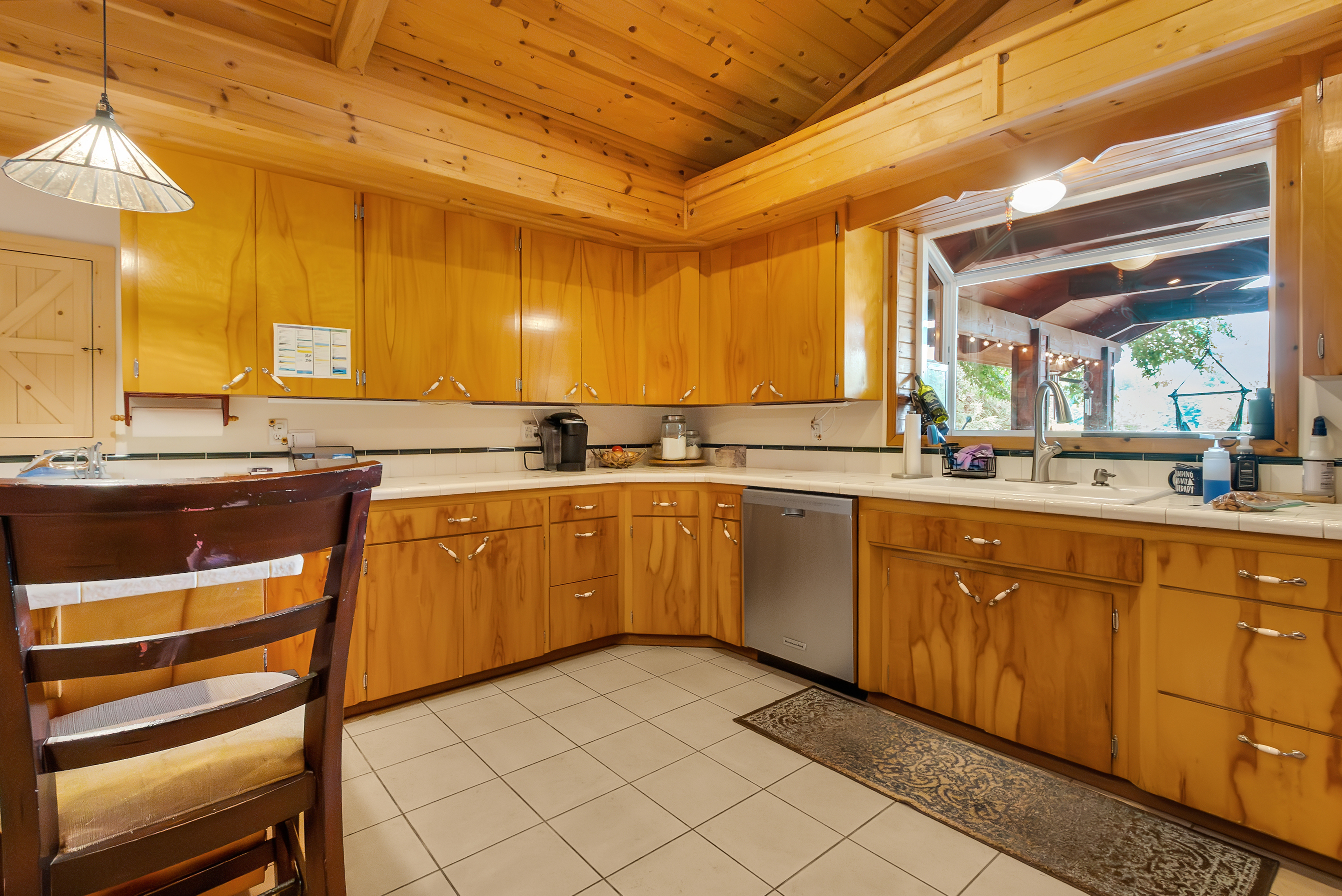This is a warm and inviting kitchen featuring wood cabinetry and a white countertop. The kitchen is well-lit with natural light coming through a window above the sink. Stainless steel appliances and a tile floor complete the space, creating a functional and aesthetically pleasing area.