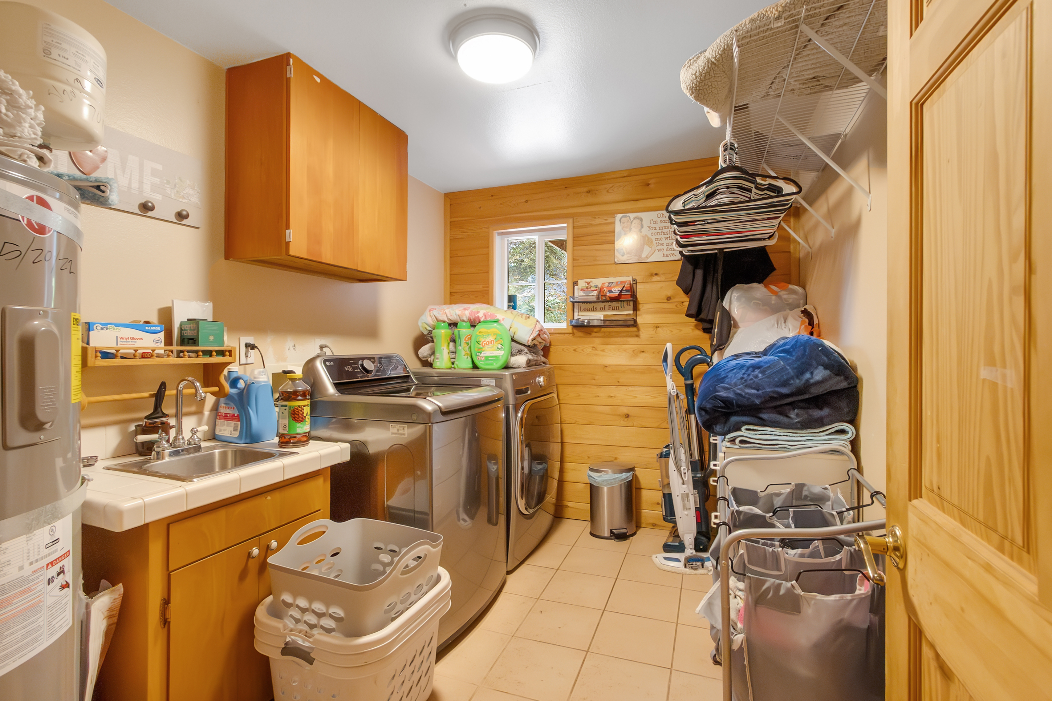 This is an interior shot of a laundry room. The room features a washer and dryer set, a sink with wooden cabinetry, and overhead storage. The walls are a mix of painted drywall and wood paneling, and the floor is tiled. The room is somewhat cluttered with laundry supplies and other items.