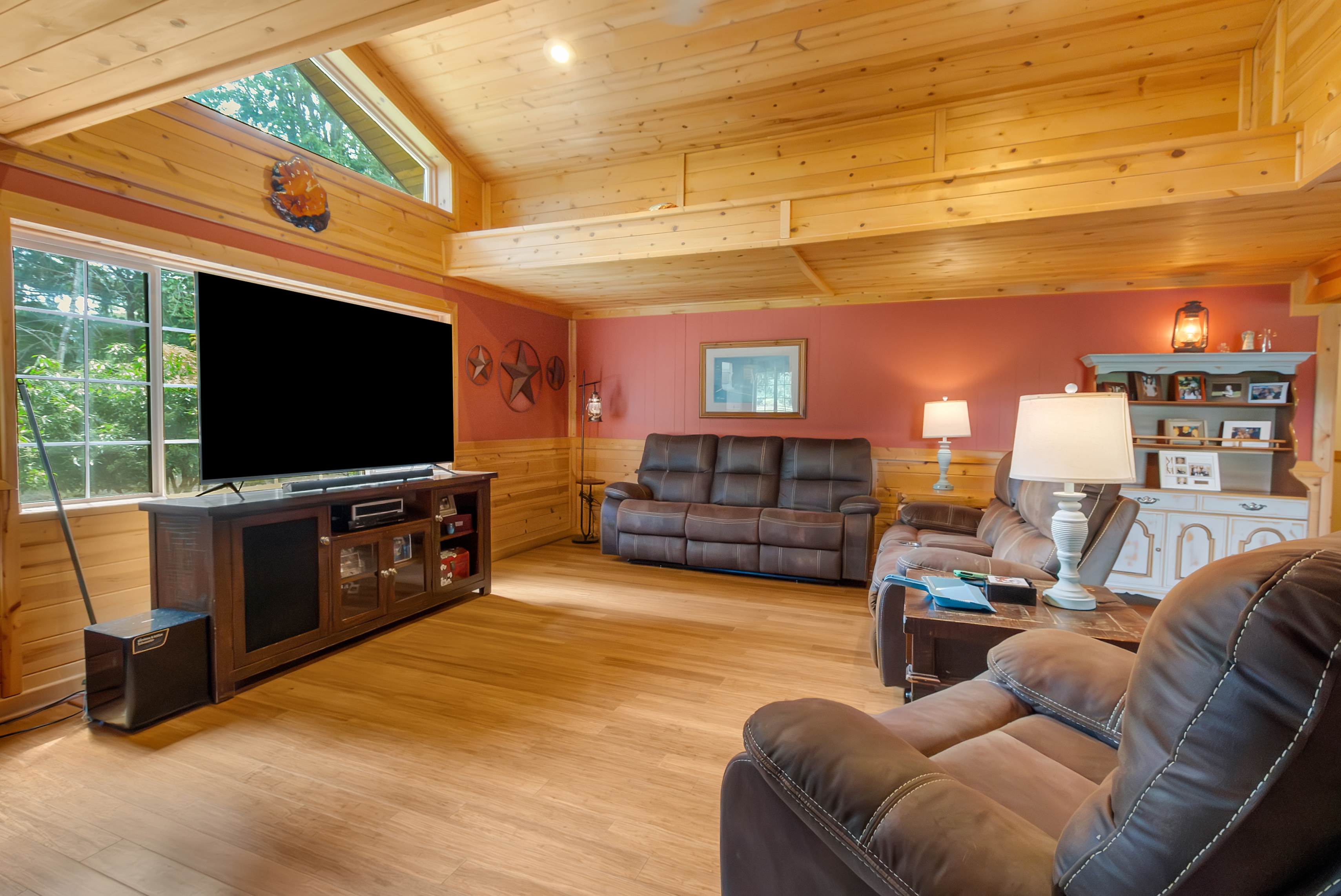 This is an interior shot of a cozy living room featuring wood-paneled walls and ceiling, creating a warm and inviting atmosphere. The room is furnished with comfortable brown leather reclining sofas and a matching loveseat, centered around a wooden entertainment center with a large television. Natural light streams in through a large window, enhancing the room's appeal.