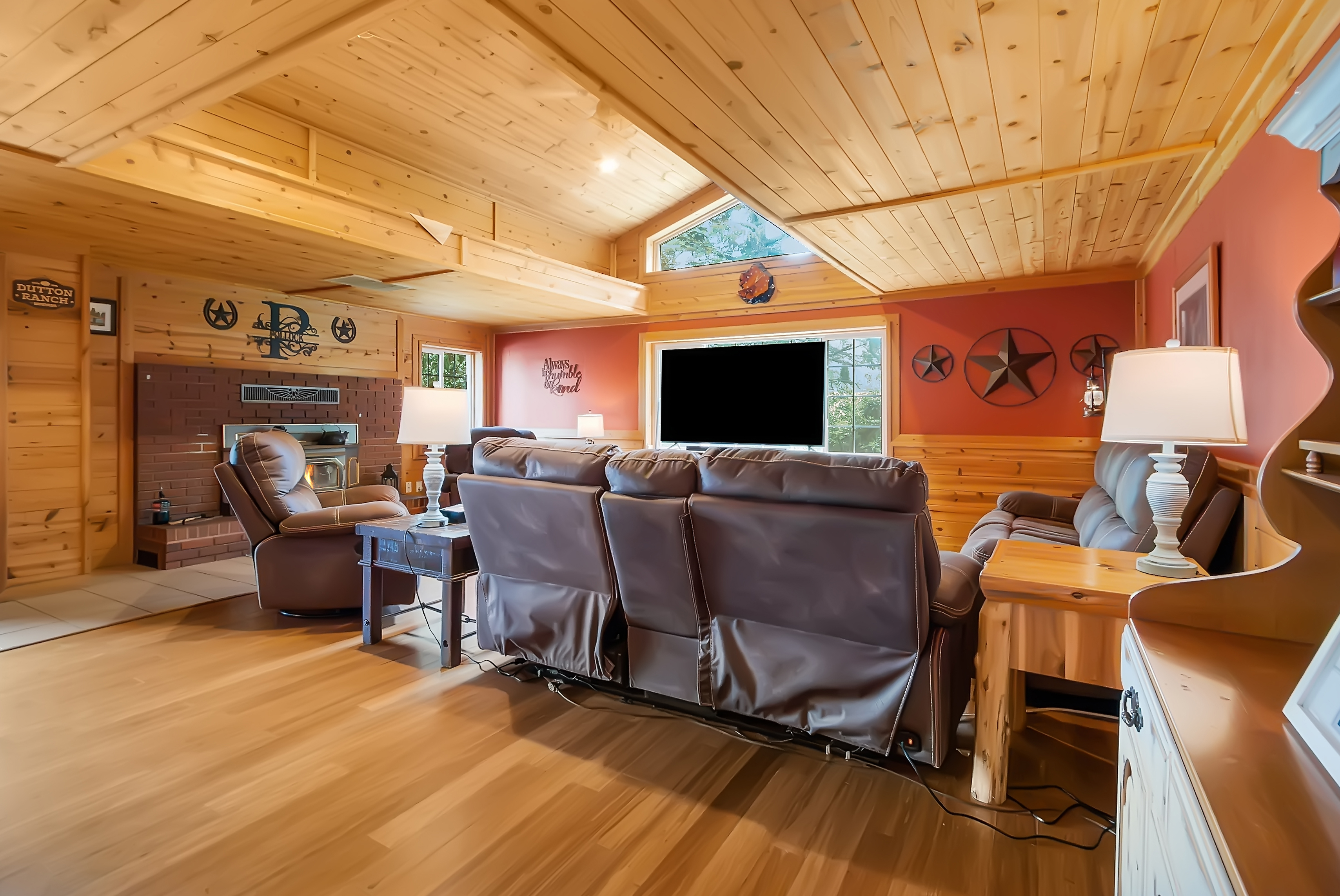 This is a warm and inviting living room featuring wood-paneled walls and ceiling, creating a cozy, rustic atmosphere. The room is furnished with comfortable leather seating, including a reclining sofa and armchair, arranged around a brick fireplace and a large television. Decorative elements like metal stars and a personalized sign add character to the space.