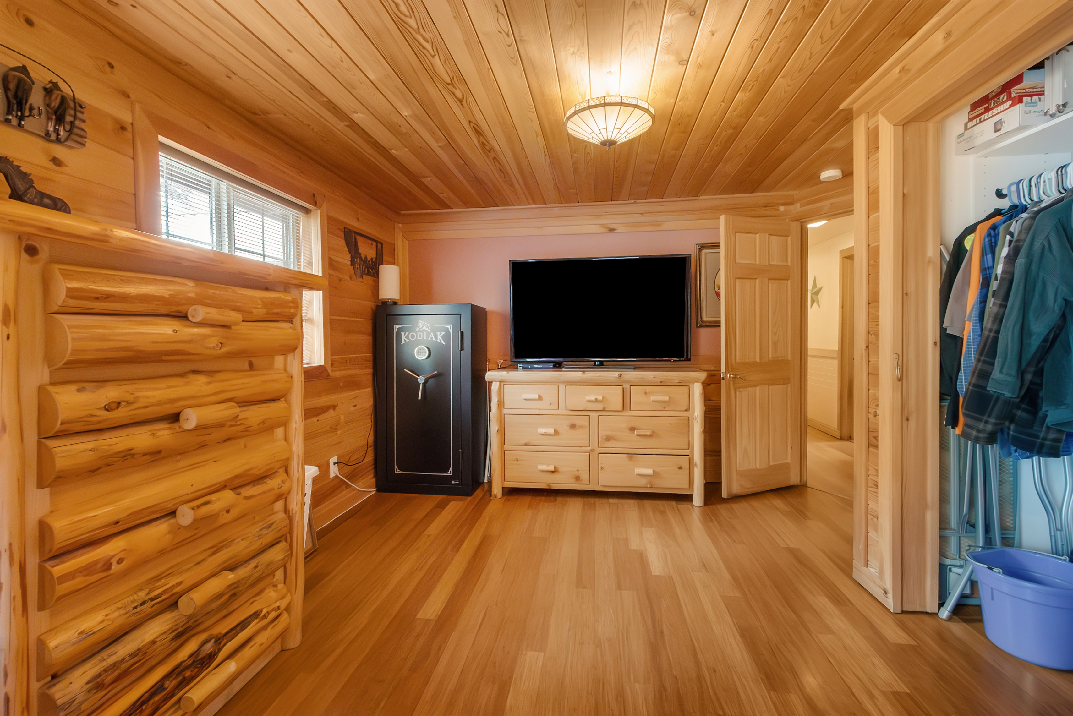 This is a cozy bedroom with wood-paneled walls, ceiling, and hardwood floors, creating a warm and inviting atmosphere. The room features a log-style dresser, a flat-screen TV on a wooden dresser, and a black safe. An open doorway leads to a hallway, and a closet is visible on the right, suggesting ample storage space.