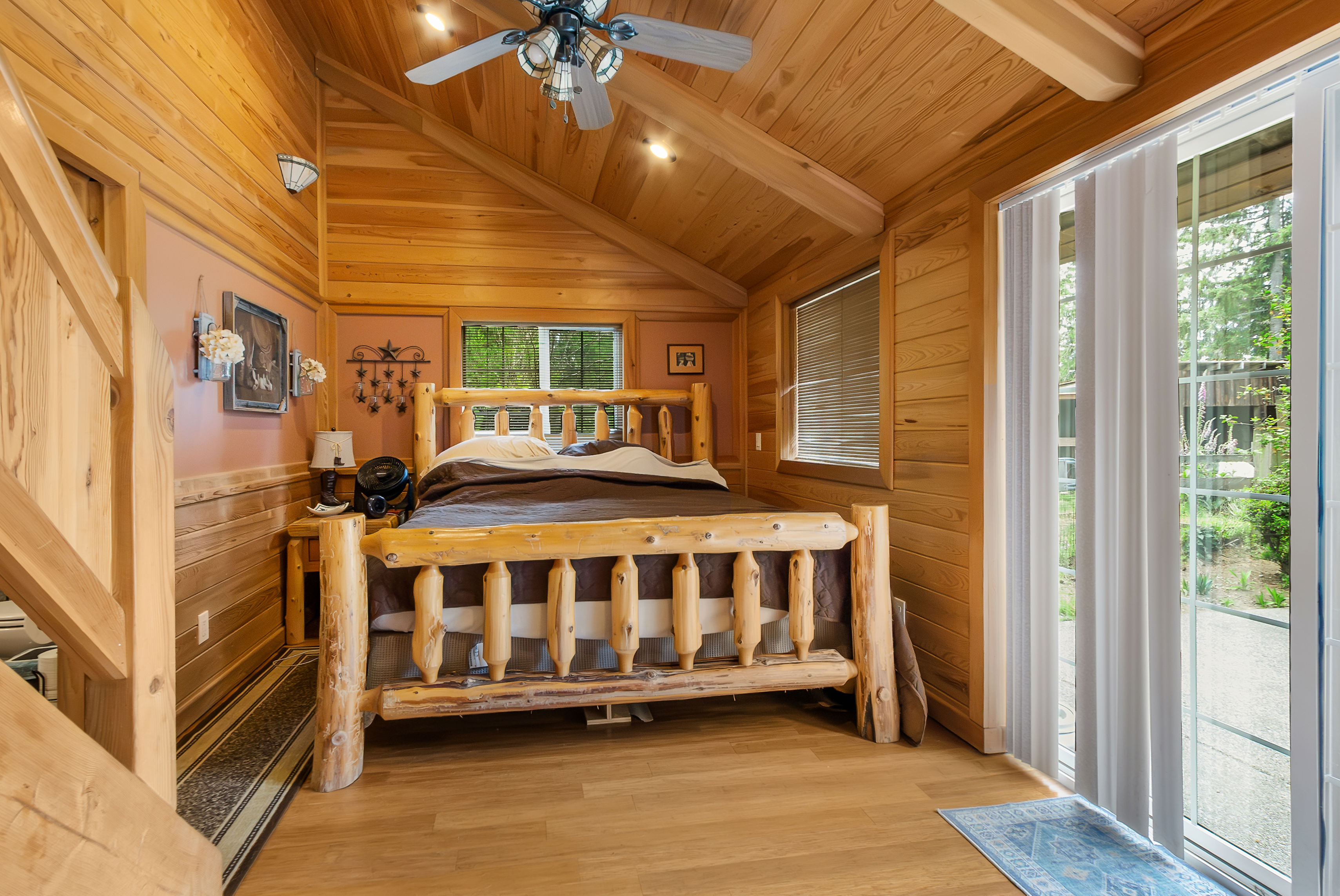 This is a cozy bedroom featuring a rustic log bed with a brown comforter. The room is paneled with wood, creating a warm and inviting atmosphere. Natural light streams in through the windows and sliding glass door, enhancing the cabin-like feel of the space.