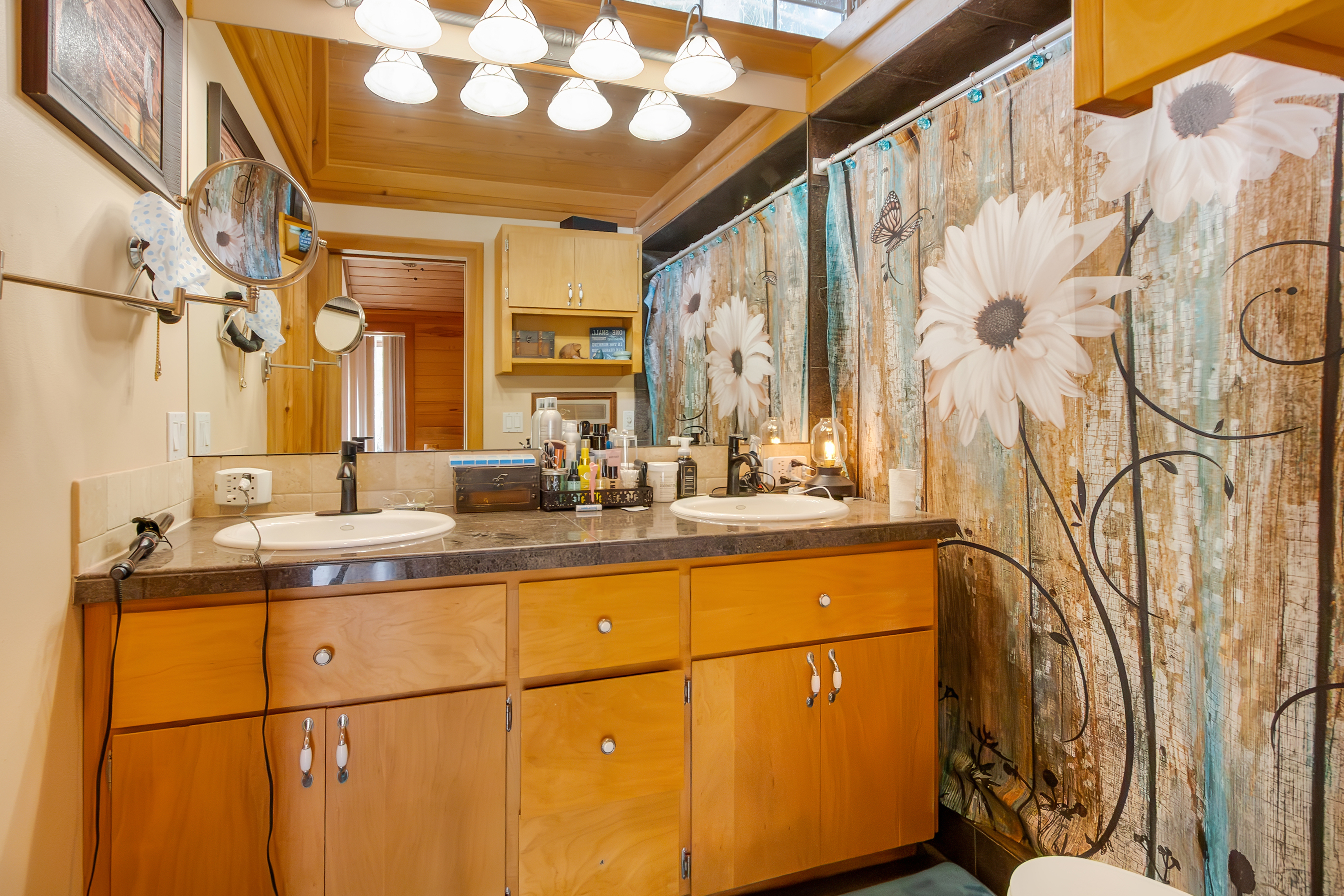 This is a bathroom featuring a double vanity with a dark countertop and light wood cabinets. Above the vanity is a large mirror with decorative lighting. The shower area is concealed by a decorative shower curtain with a floral pattern, adding a touch of whimsy to the space.