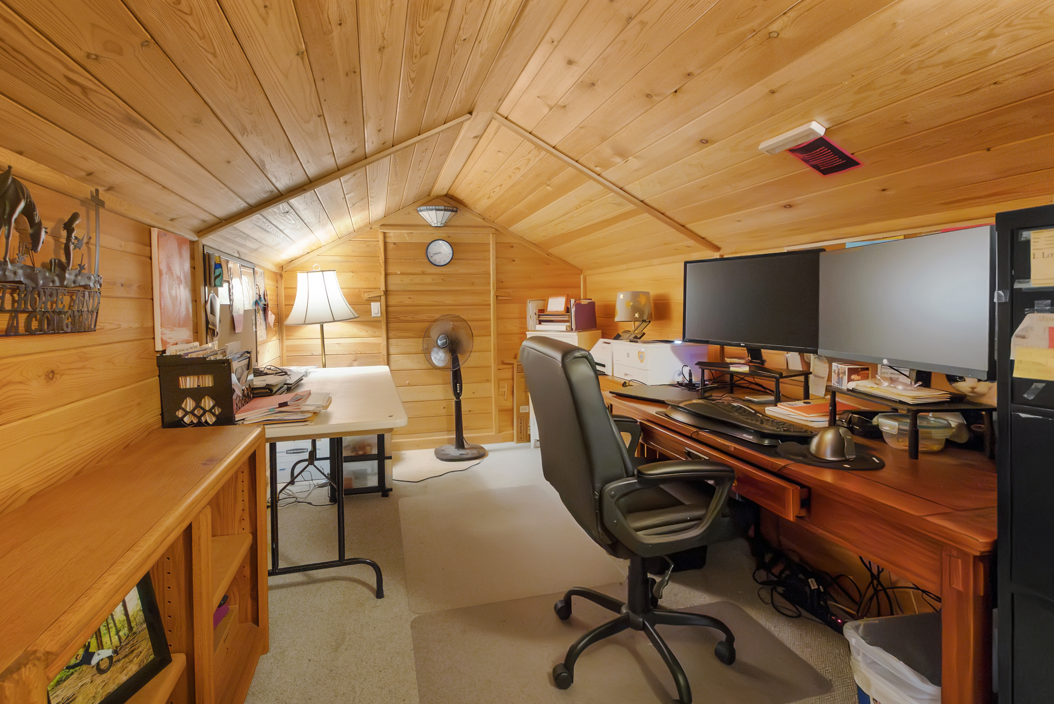 This is an interior shot of a cozy office or study space, likely located in an attic due to the sloped wooden ceiling and walls. The room features a wooden desk with dual monitors, an ergonomic office chair, a standing fan, and a secondary desk with a lamp. The overall impression is a warm and functional workspace.