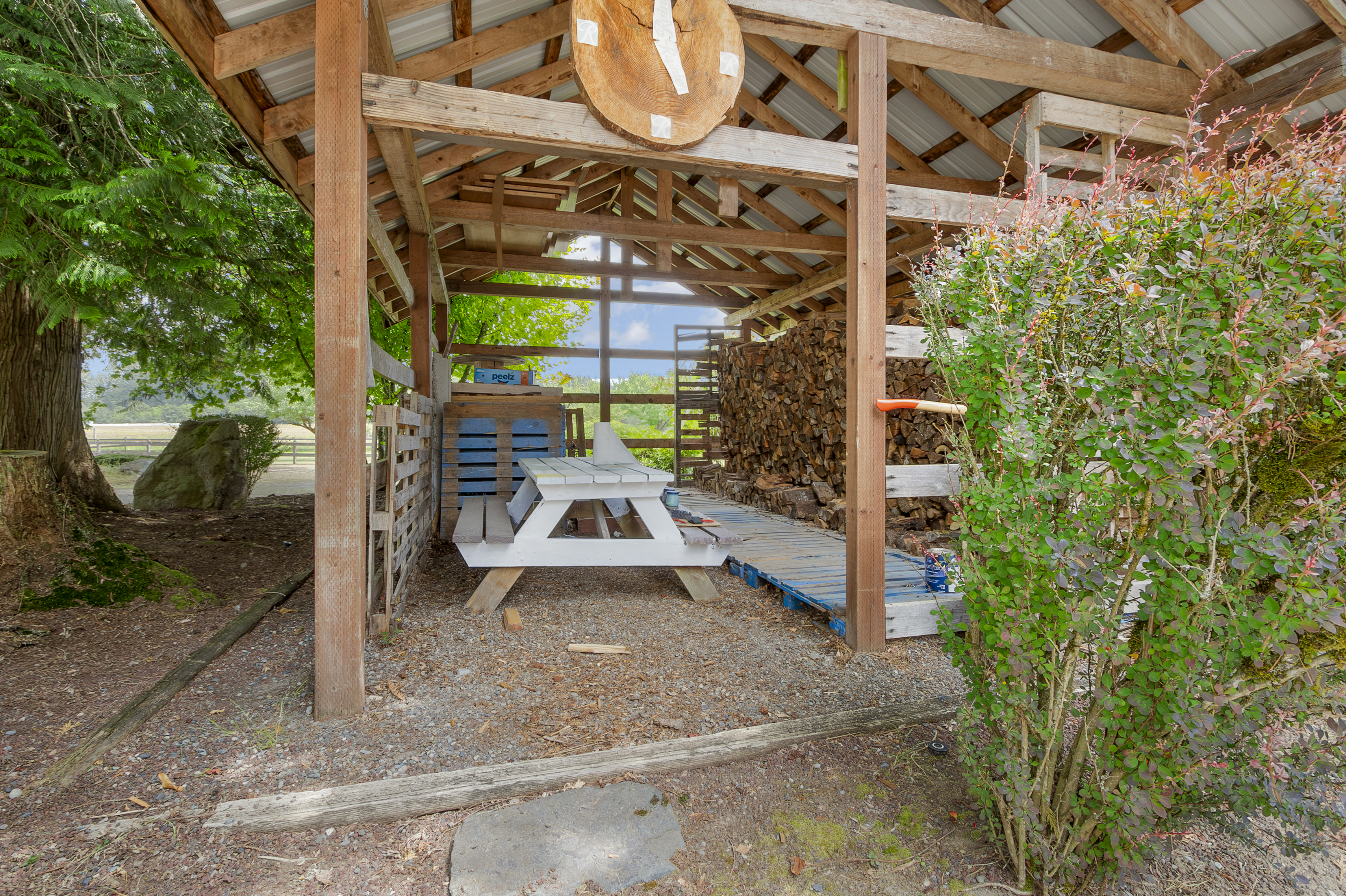 This image showcases a rustic outdoor shelter, possibly a wood storage area or picnic spot, featuring a wooden structure with a metal roof. Inside, there's a white picnic table and stacks of firewood, creating a functional and charming space. The surrounding yard includes gravel, a stone path, and a vibrant bush, adding to the natural ambiance.