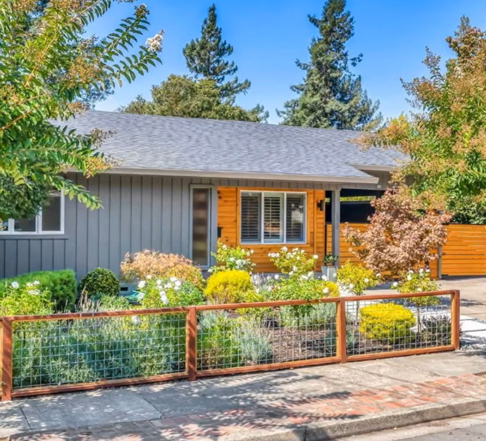 This charming modern home features a striking combination of grey vertical siding and natural wood paneling, which adds significant curb appeal. A wire-mesh fence with wood framing defines the front yard, which is landscaped with a mix of lush green shrubs and flowering plants. The architectural style reads as a contemporary update to a classic ranch design, presenting a warm and inviting front facade.