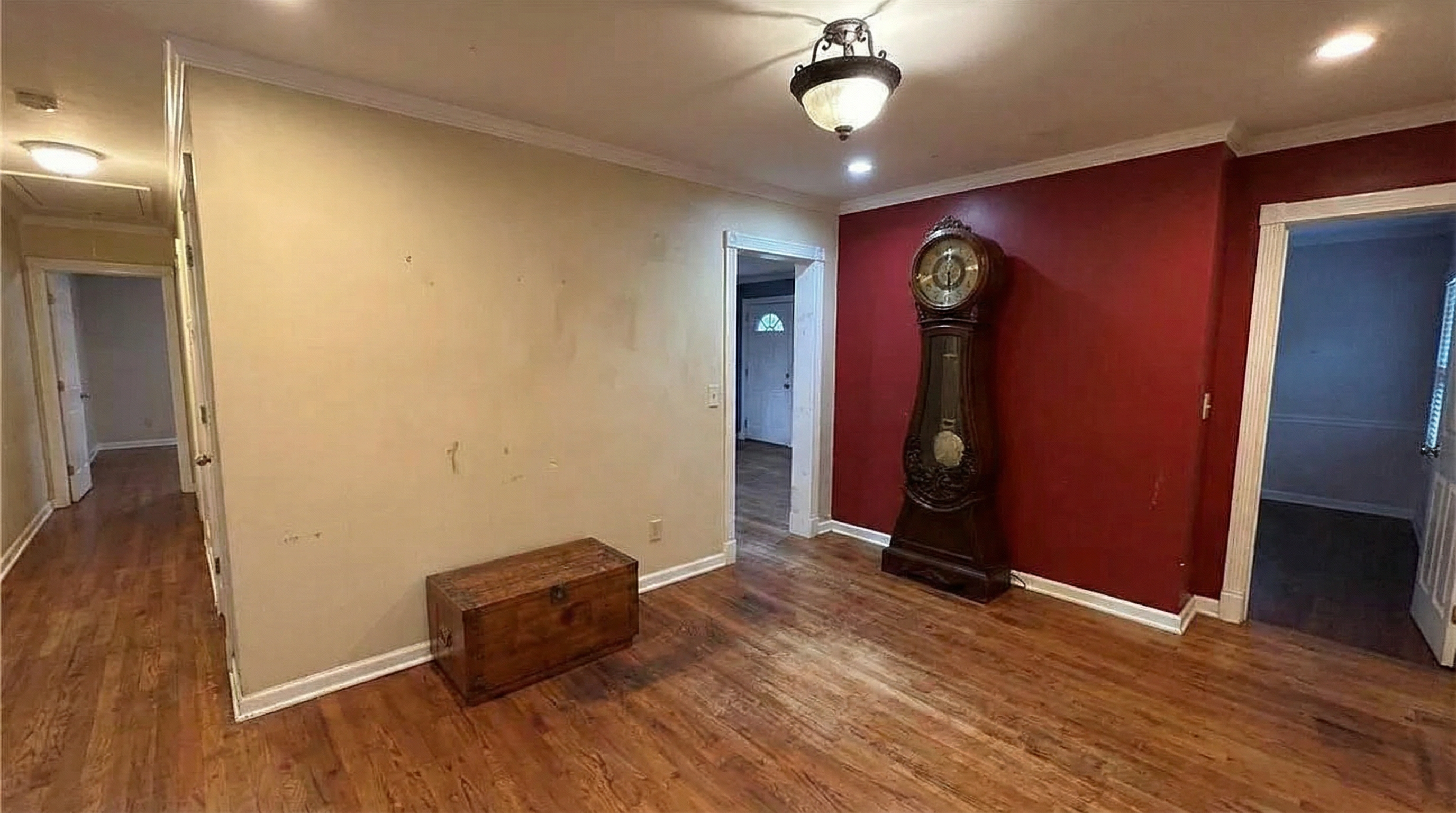 The image showcases a living room with hardwood flooring, a light-colored wall, and a contrasting maroon accent wall. A vintage-looking grandfather clock stands against the maroon wall, adding a touch of character. A wooden chest sits in the room providing potential for storage or decor.