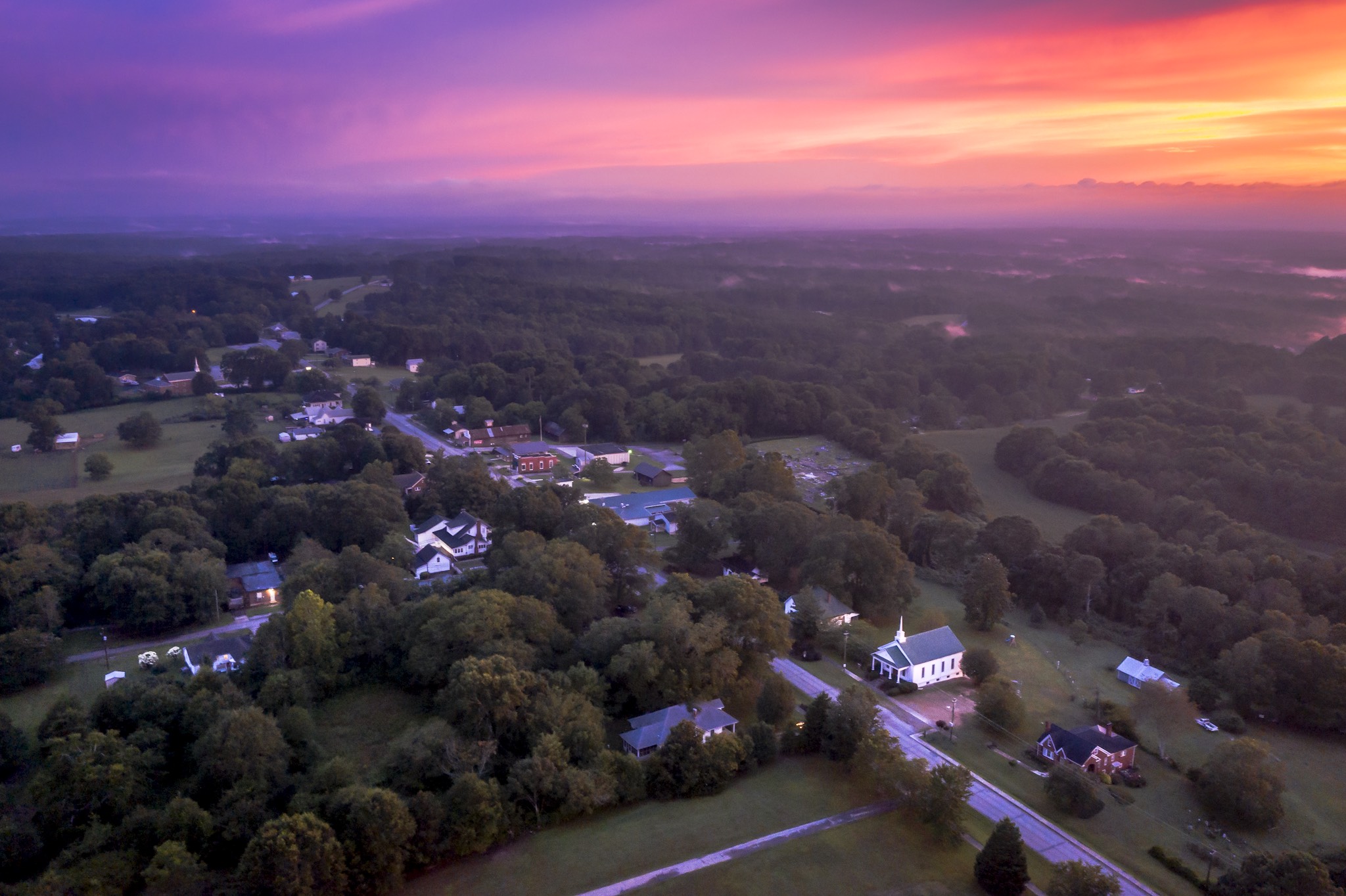 This aerial view showcases a quaint, rural setting at dusk, featuring a mix of residential properties, forested areas, and a distinctive white church with steeple. The lighting from the sunset paints the sky in shades of purple and orange, creating a picturesque and serene ambiance, that would appeal to buyers seeking a peaceful community.