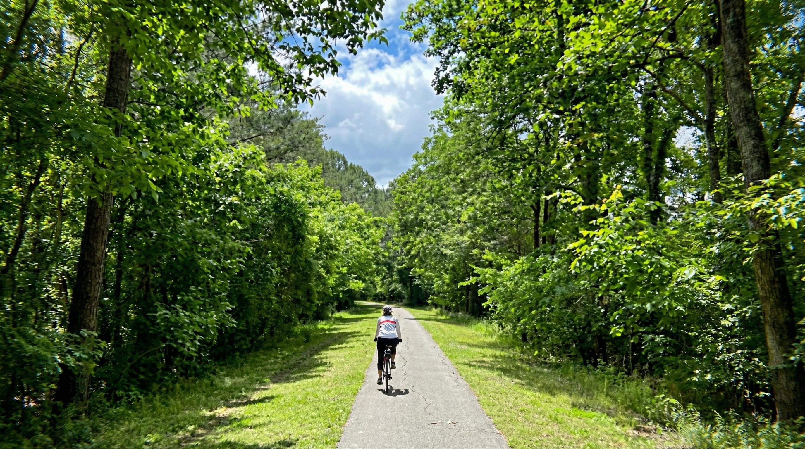The image showcases a scenic paved path, likely part of a community trail or park. The path is framed by lush greenery, creating an inviting atmosphere for walking, biking, or other recreational activities. A cyclist is visible on the path further enhancing the amenity's appeal.