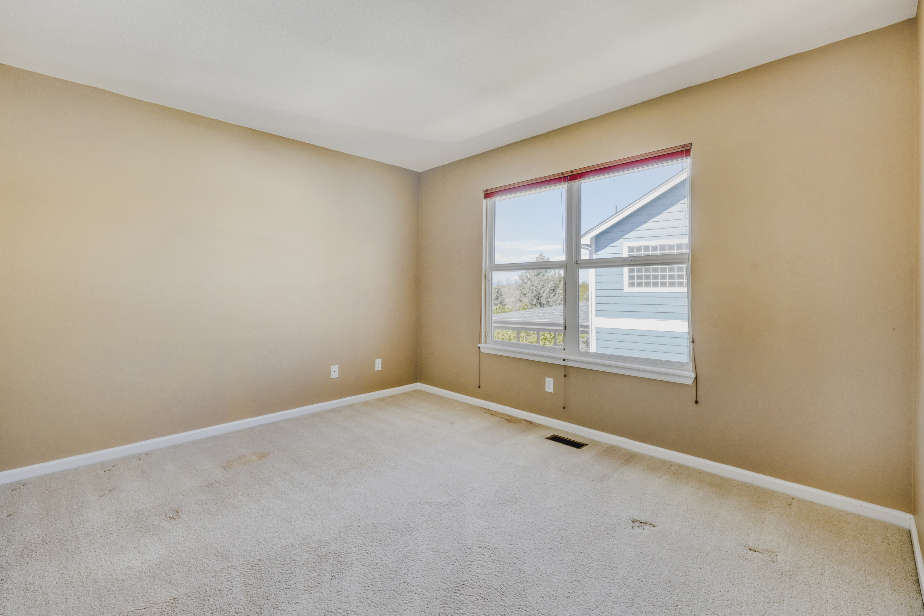 This is a quiet, empty bedroom featuring neutral-toned walls and beige carpeting throughout. The space is highlighted by a large window that invites natural light, providing a view of the neighboring property. It presents a versatile, blank canvas ready for personalization and design.