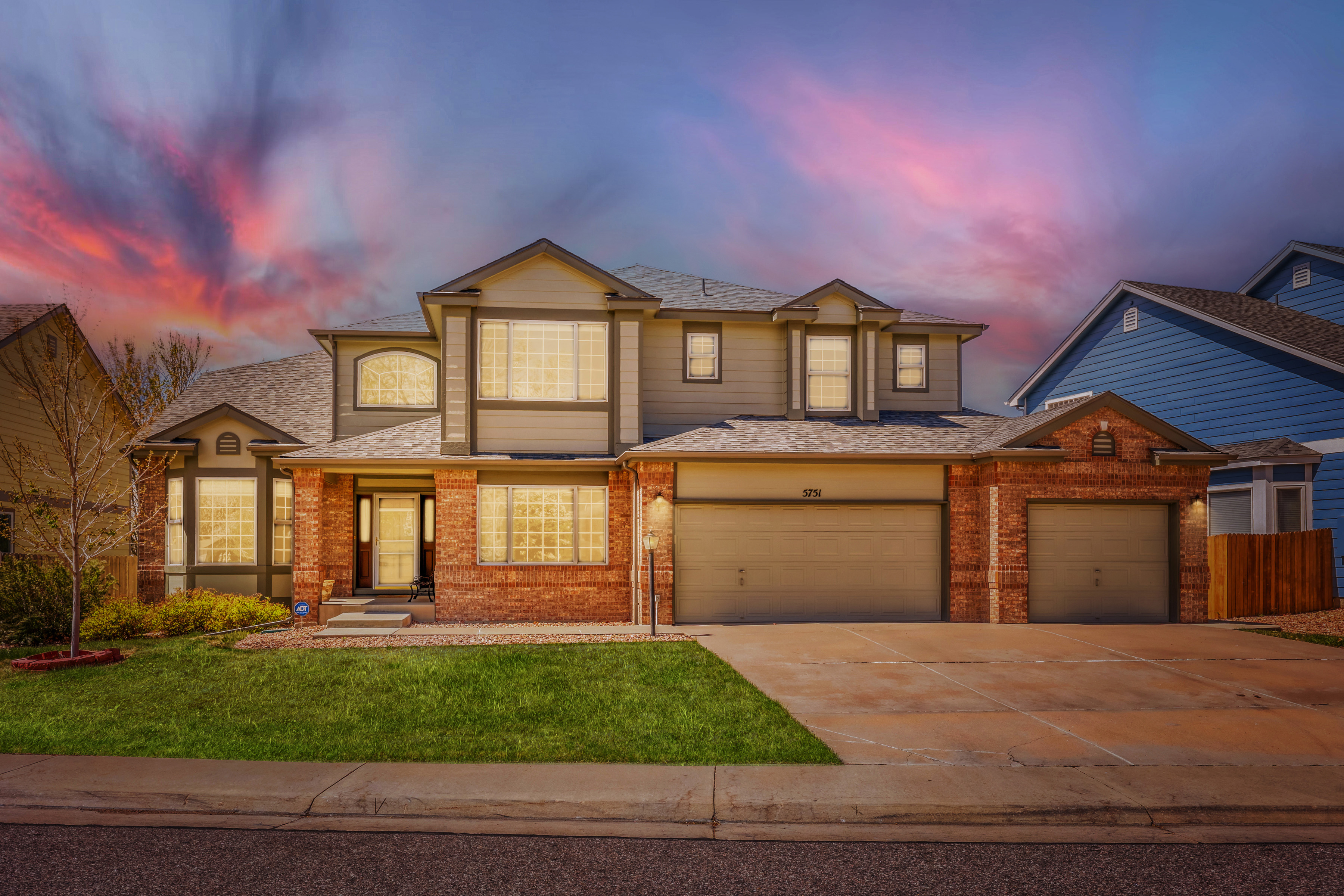 This is a beautiful two-story residential home with a classic blend of brick and siding architecture. The home features a spacious three-car garage, a well-manicured front lawn, and is captured during a dramatic sunset that enhances its curb appeal. The welcoming entryway and invitingly lit interior windows suggest a warm and comfortable living space.