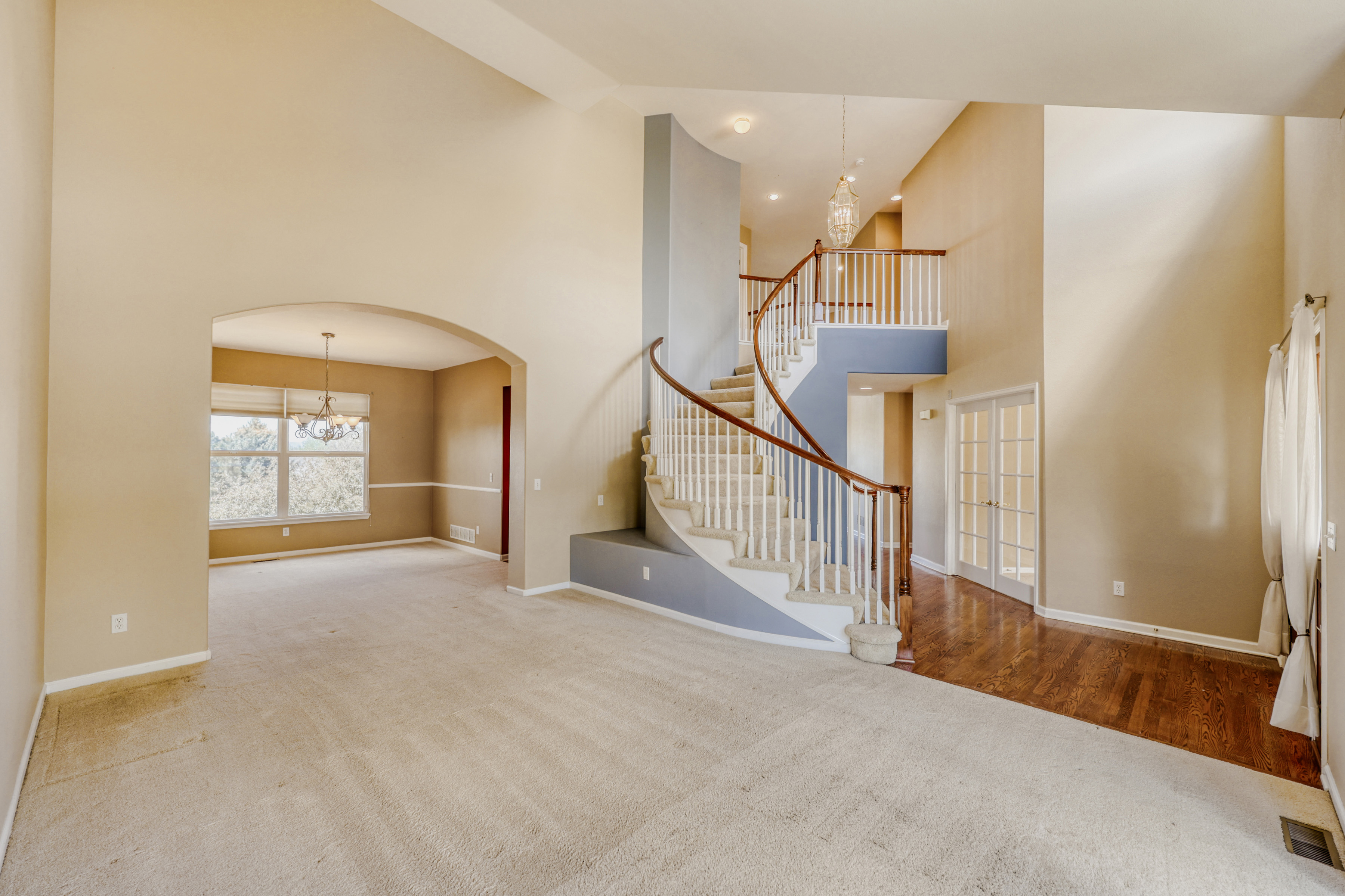 This spacious foyer features a grand curved staircase as its focal point, complemented by tall ceilings and neutral wall tones. The area combines carpeted flooring with rich hardwood accents near the entryway and features an elegant chandelier hanging above the stairs. The open-concept design transitions seamlessly into a formal dining area, creating an inviting and airy atmosphere.