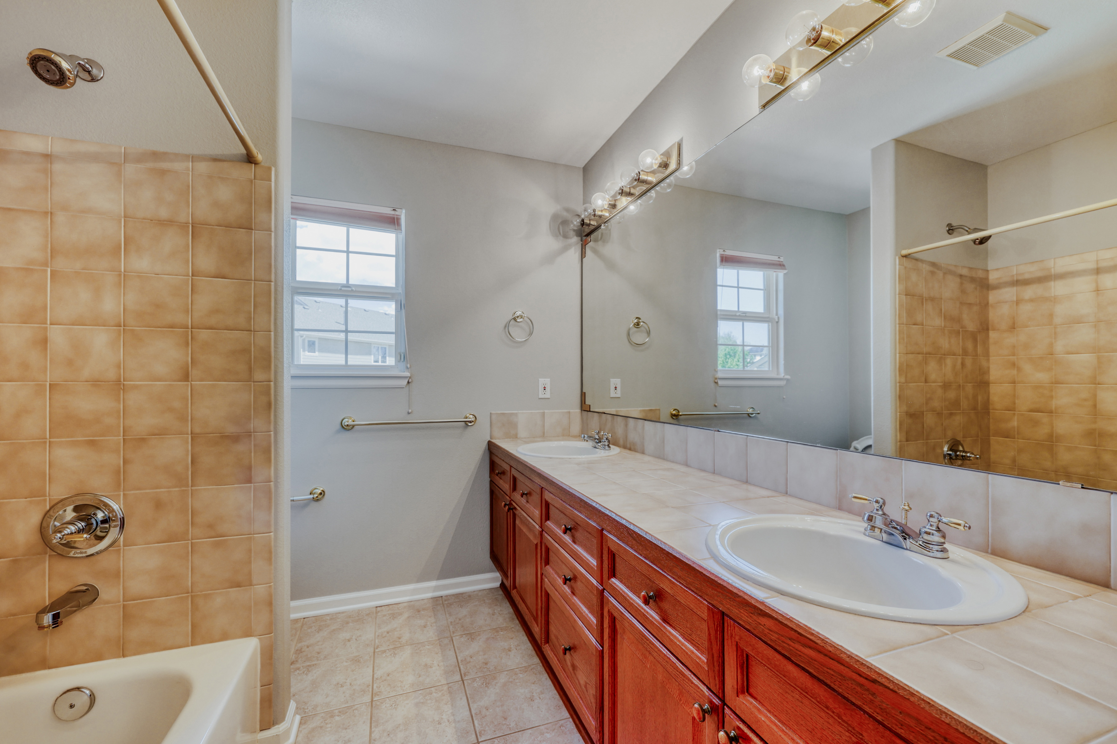 This primary bathroom features a long, dual-sink vanity with dark wood cabinetry and tiled countertops, offering ample storage space. A large wall-mounted mirror enhances the sense of space, while the adjacent tub and shower combination is finished with warm-toned ceramic wall tiles. The neutral wall color and practical layout provide a functional and traditional aesthetic suitable for daily use.