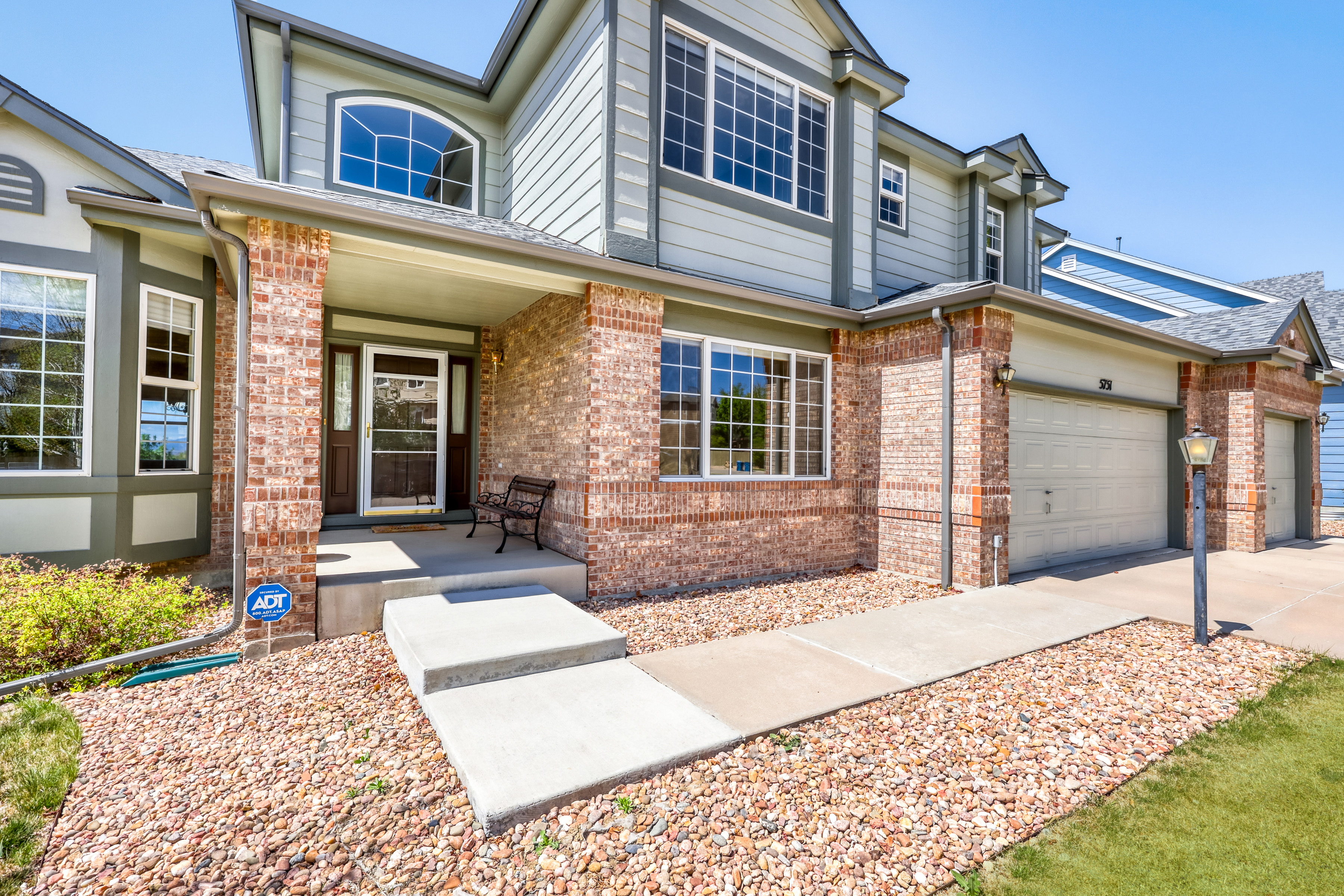 This appealing two-story suburban home features a classic blend of red brick and light-colored siding, giving it a charming, traditional aesthetic. The front entrance is highlighted by a cozy porch area and a walkway leading to a multi-car garage, while the landscaping consists of tidy rock beds and an established lawn. It is a well-maintained property that presents a welcoming and curb-appealing exterior.