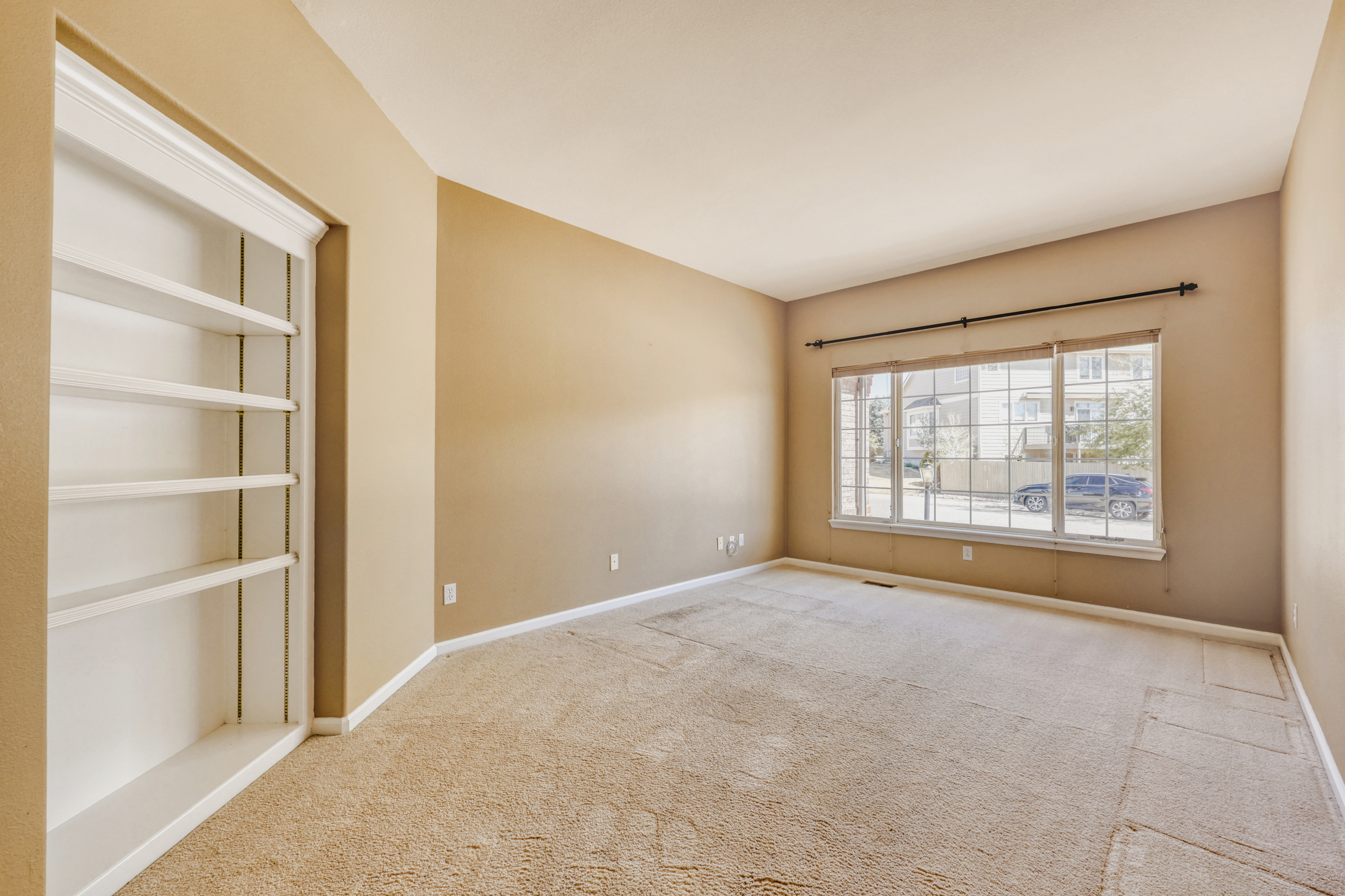 This empty room features neutral-toned walls, beige carpeting, and a built-in white bookshelf along one wall. A large window provides ample natural light and a view of the exterior, making it a versatile space perfect for a home office, den, or study.