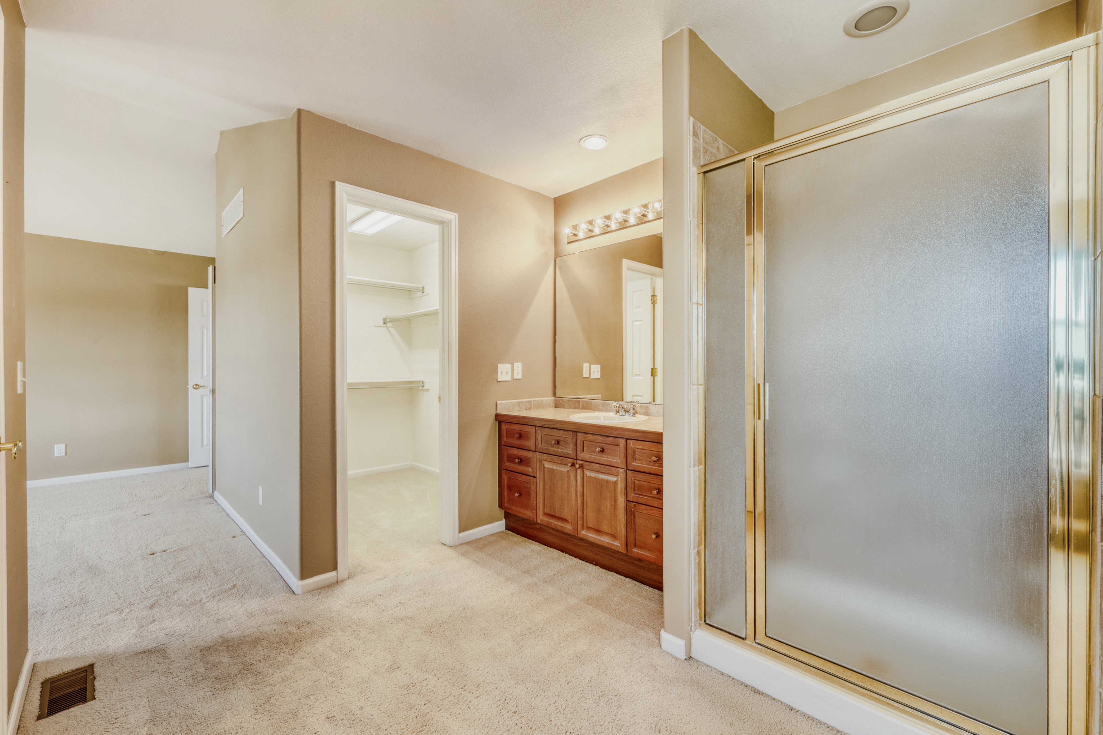 This primary bathroom features a warm, neutral color palette with beige walls and soft carpeting throughout. The space includes a functional vanity with wood cabinetry, a large mirrored wall, and a convenient glass-enclosed shower stall. An adjacent walk-in closet adds extra storage capacity, completing this practical and accessible residential suite.