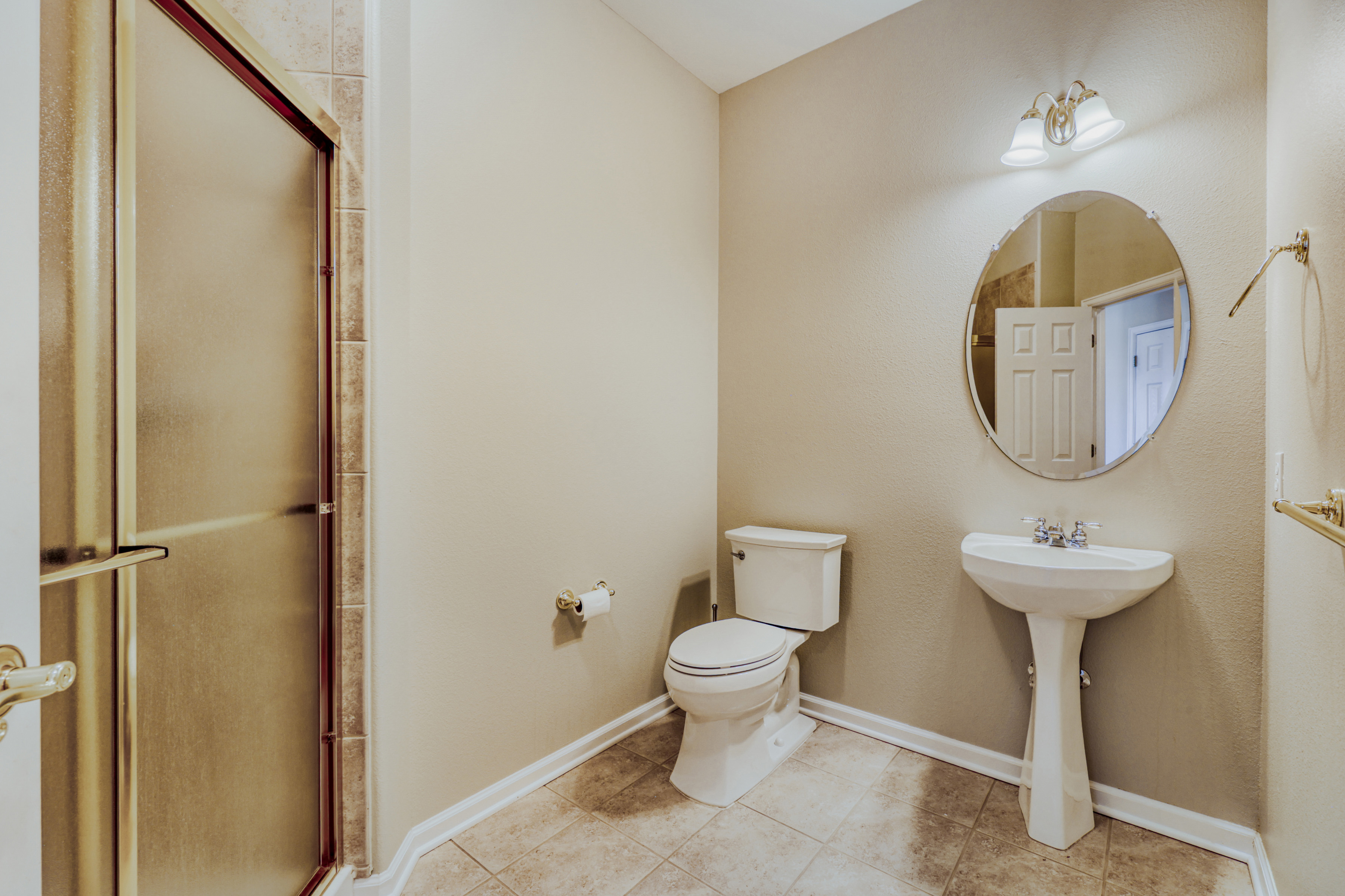 This clean and functional bathroom features a pedestal sink, a standard white toilet, and a tiled shower enclosure with a glass door. The space is finished in neutral-toned paint with ceramic floor tiles and gold-toned hardware, offering a simple and accessible layout.