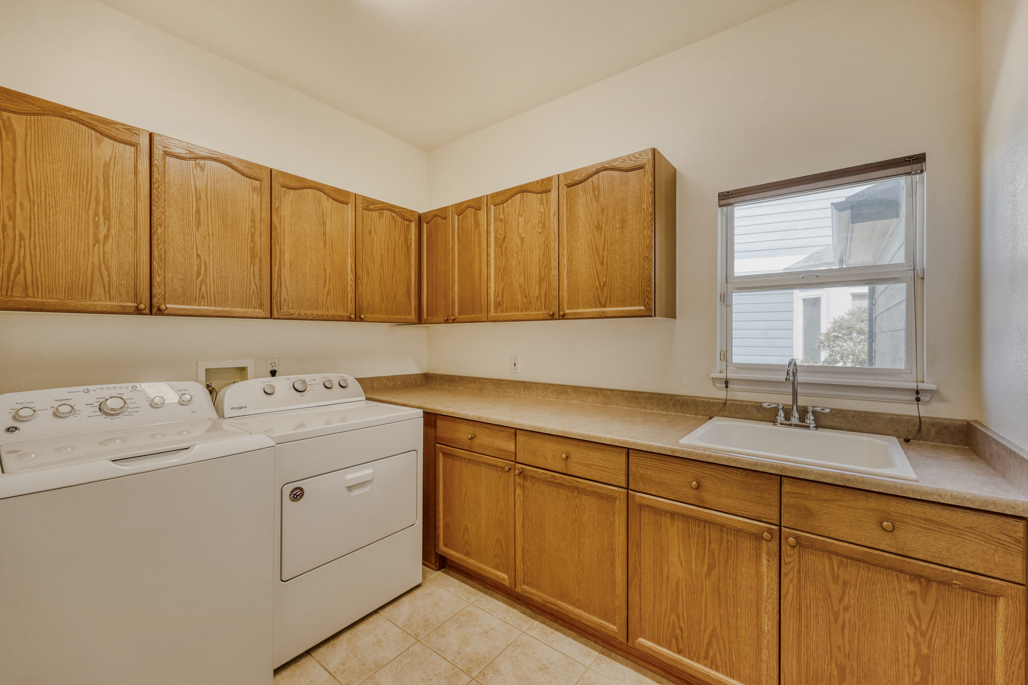 This functional laundry room features classic light oak cabinetry providing abundant storage space above and below the laminate countertop. It is equipped with a clean white utility sink and side-by-side washer and dryer units, making it a highly practical workspace. A window above the sink illuminates the area with natural light, contributing to the room's tidy and traditional aesthetic.