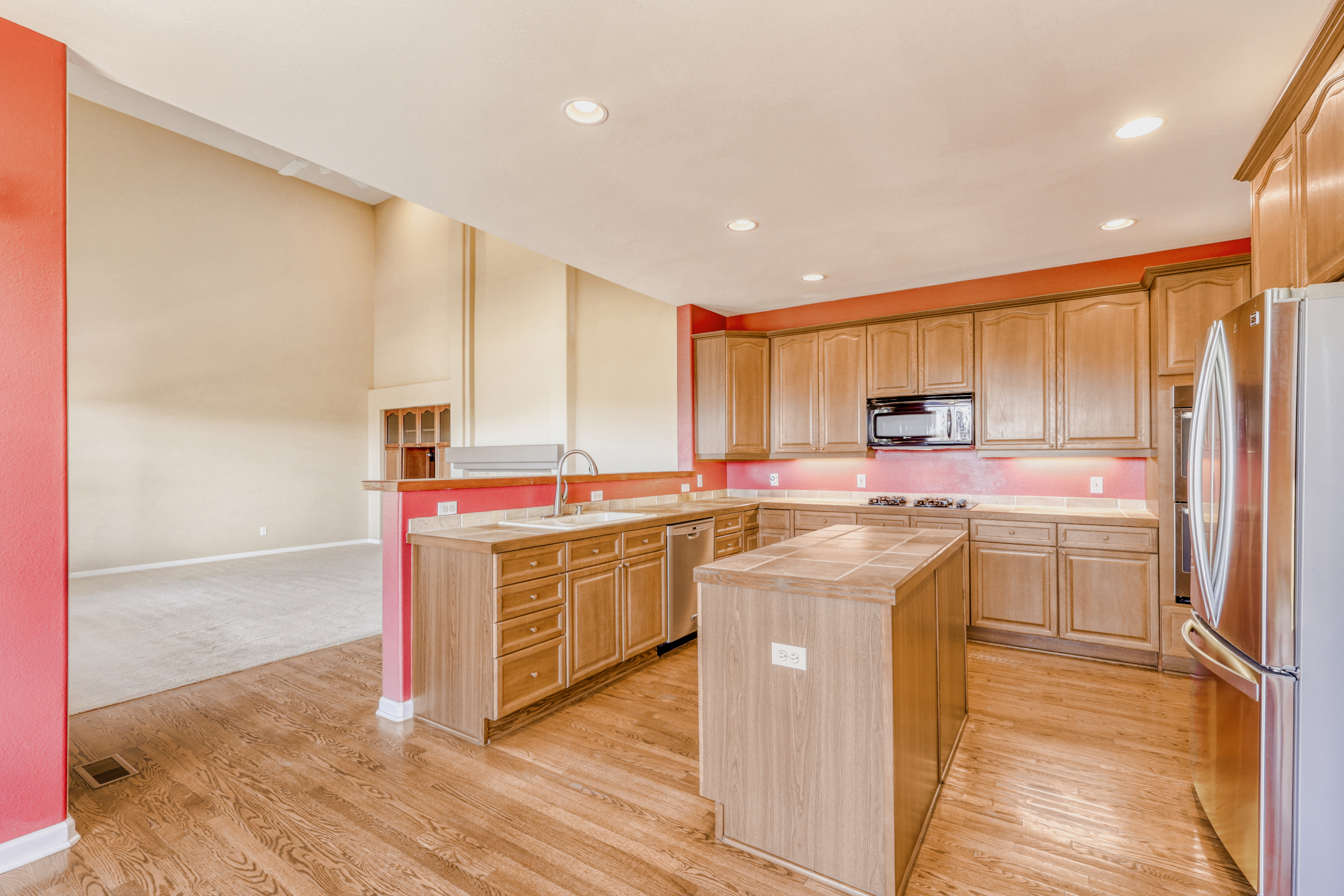 This kitchen features wood-tone cabinetry, tiled countertops, and stainless steel appliances, including a built-in microwave and refrigerator. A central kitchen island provides extra workspace, while the open-concept layout connects to a spacious adjacent living area with high, vaulted ceilings. The warm red accent walls and hardwood flooring create a distinctive and inviting aesthetic throughout the space.