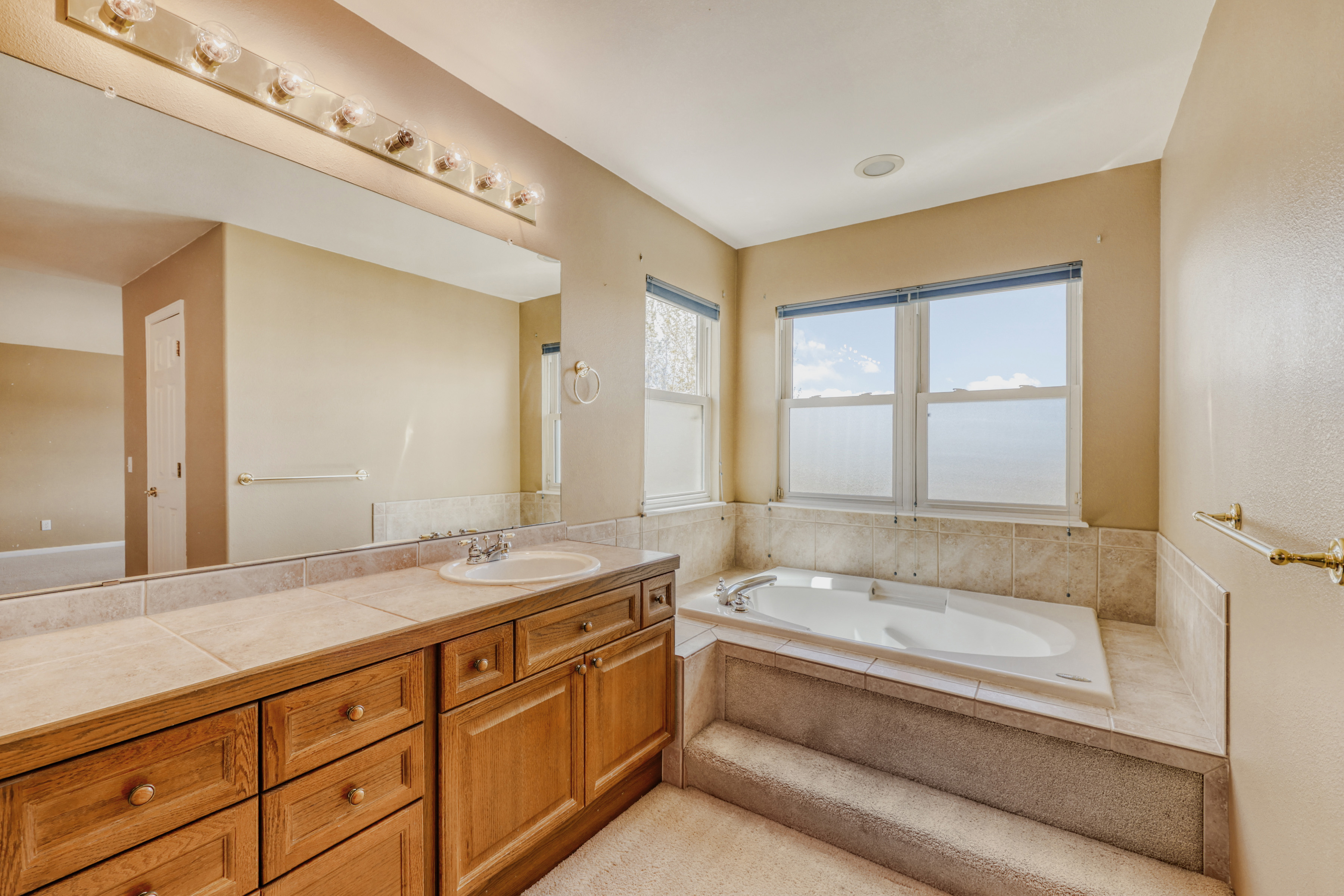 This primary bathroom features a warm, traditional aesthetic with oak cabinetry and neutral beige wall tones. It is highlighted by a deep soaking tub set within a tiled platform, a dual-drawer vanity with a tiled countertop, and a large wall-to-wall mirror. The space is well-lit by a classic multi-bulb vanity light fixture and natural light streaming through the corner windows.