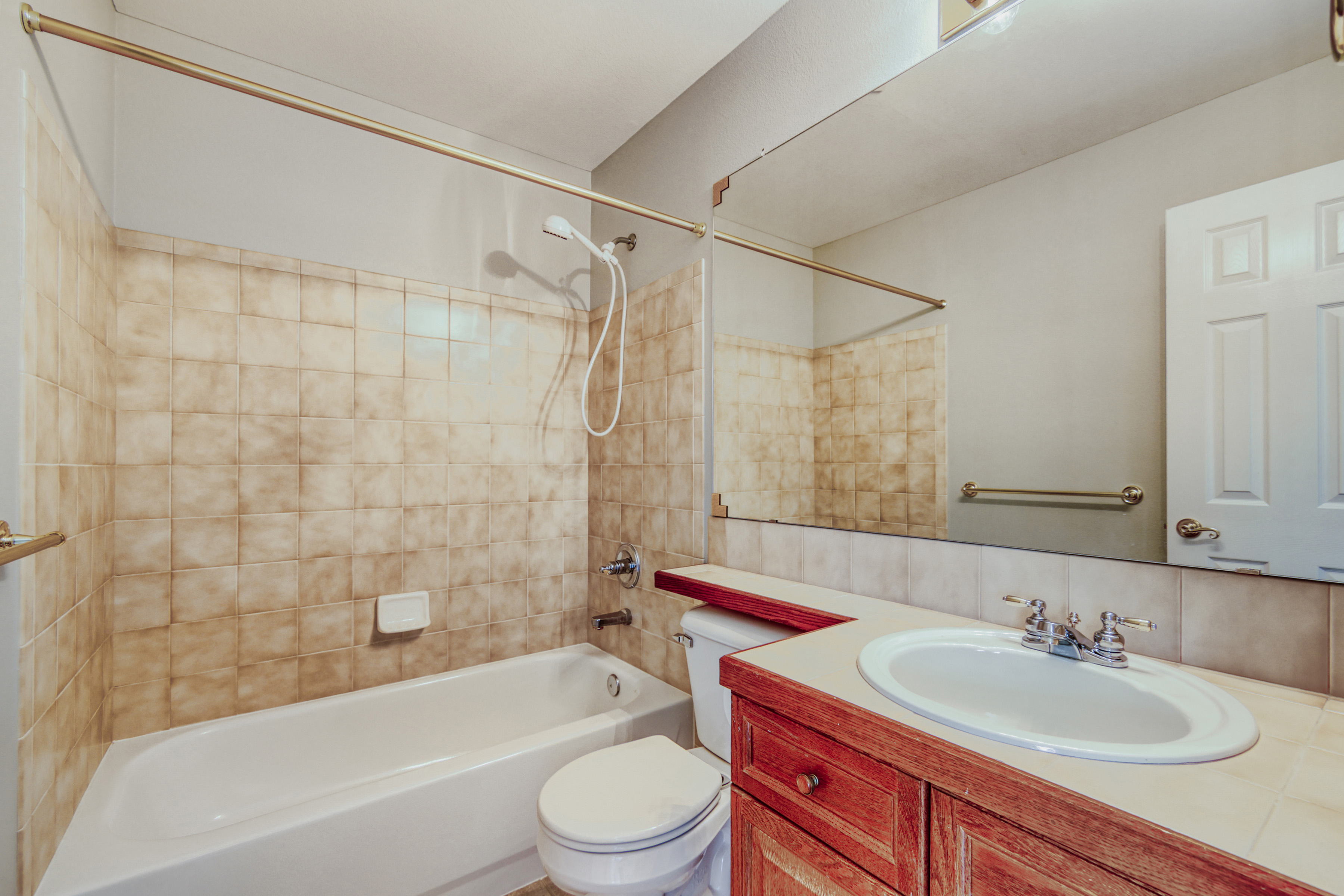 This functional bathroom features a tiled tub-shower combination with a handheld sprayer and a standard vanity with a wood-tone cabinet. The room is accented by warm, neutral beige tiling and brass-finished hardware, including the shower rod and faucet fixtures. A large wall mirror above the vanity enhances the sense of space in this clean, traditional-style bathroom.
