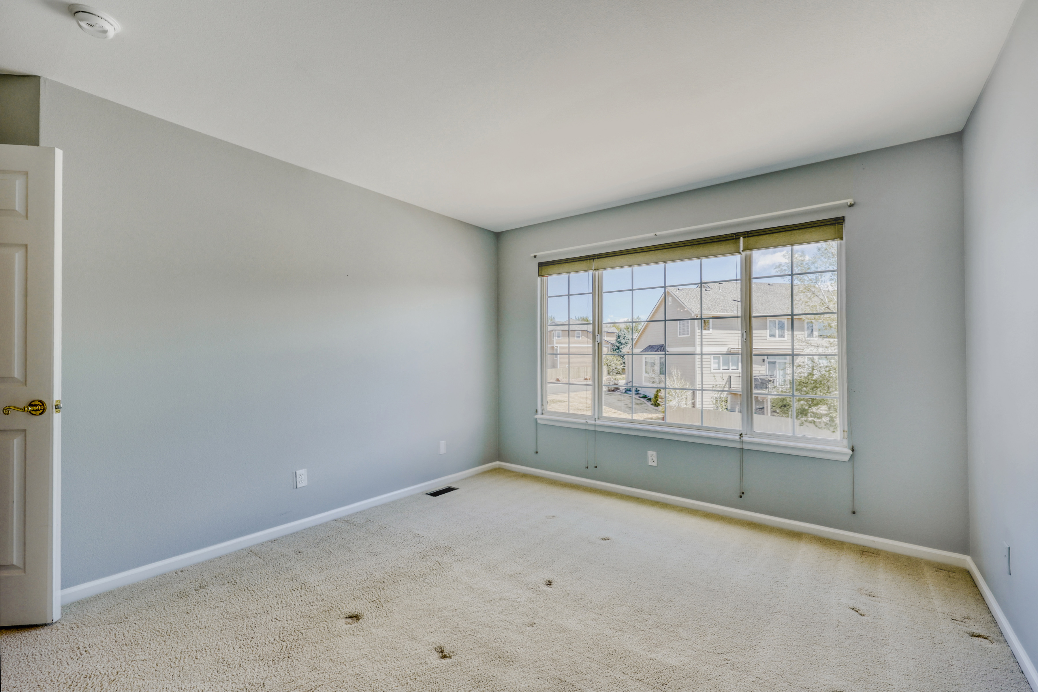 This is a clean, vacant bedroom featuring neutral-colored walls, white baseboard trim, and wall-to-wall carpeting. A large window provides ample natural light and a view of the neighboring house, while a single door provides access from the hallway. The space offers a blank canvas, making it ideal for a guest room, home office, or nursery.