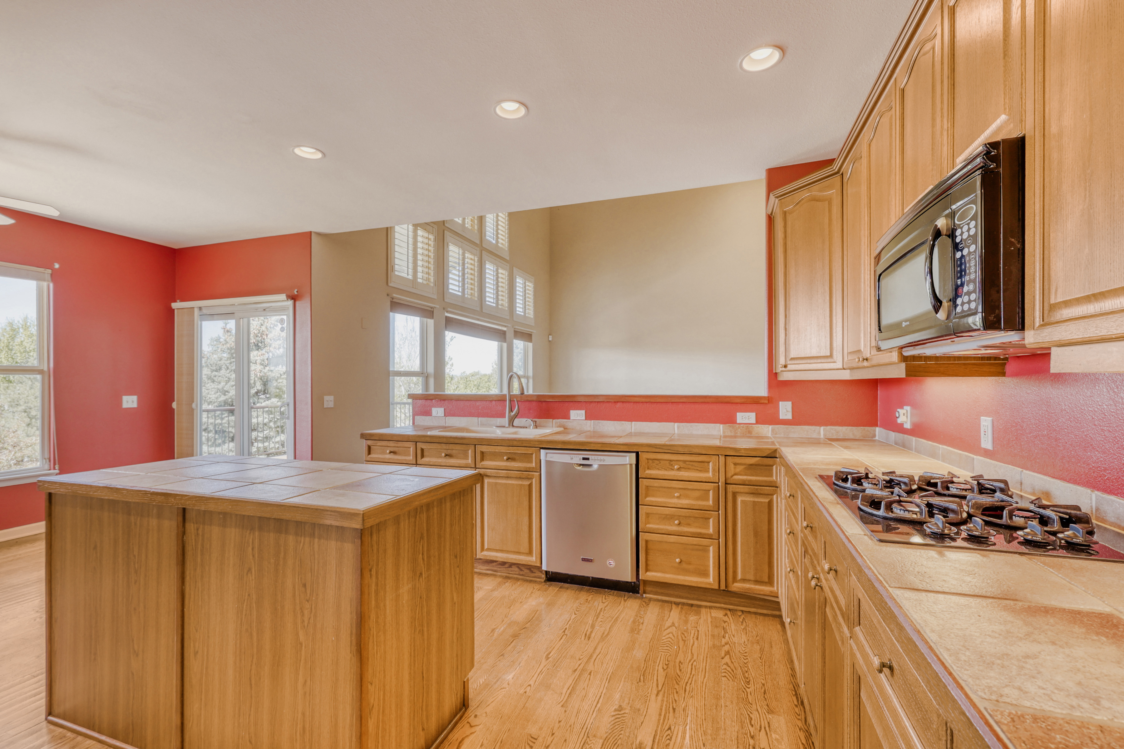 This kitchen features classic wooden cabinetry, tiled countertops, and an integrated stainless-steel dishwasher. The space is anchored by a large matching kitchen island and accented by vibrant red walls, creating a warm, traditional aesthetic. Natural light streams in from the adjacent windows, highlighting the wood flooring and open layout.