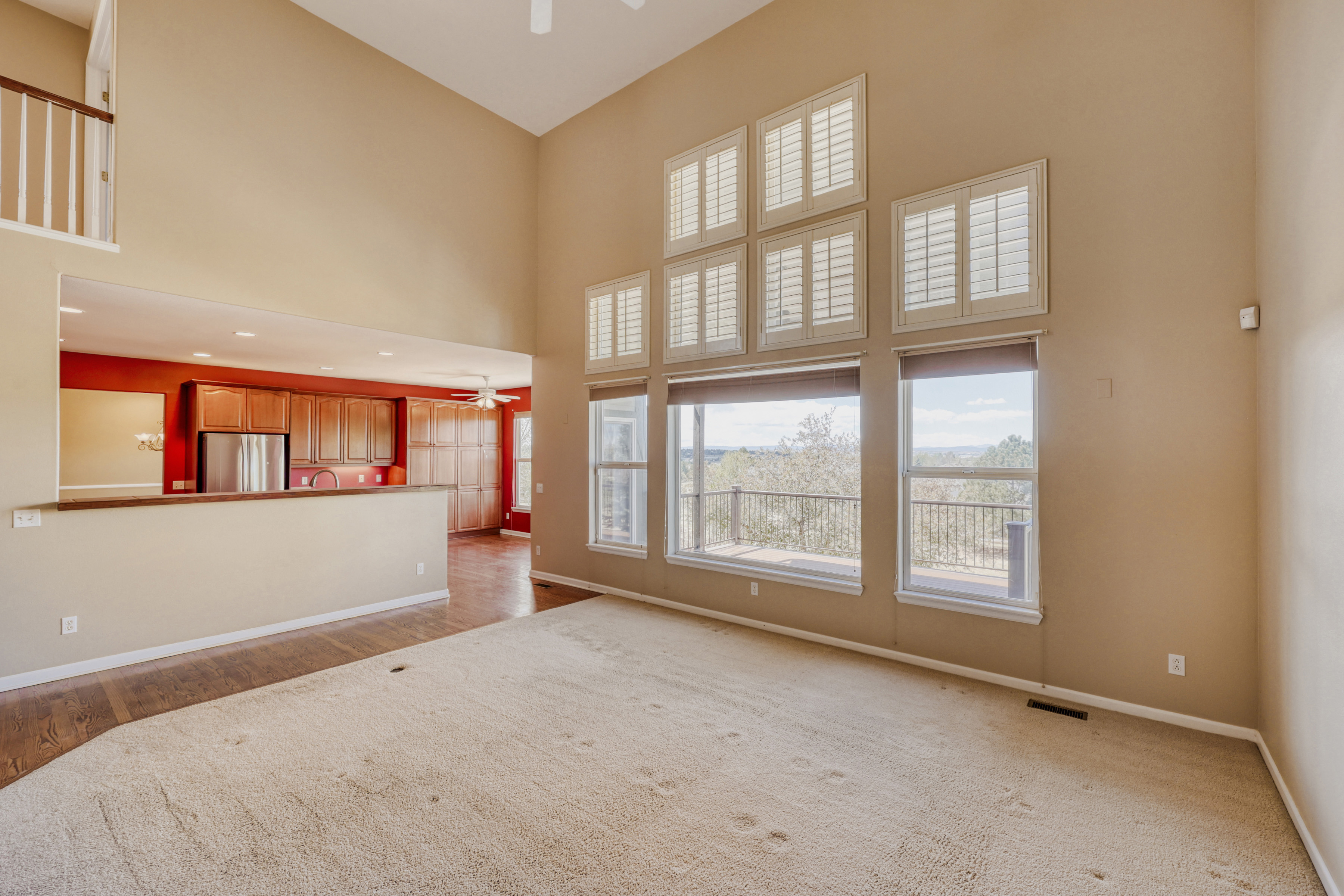 This living room is characterized by its dramatic vaulted ceilings and a expansive wall of windows that provides abundant natural light and scenic views. The layout flows openly into a red-accented kitchen, creating a spacious and connected atmosphere. The neutral-toned carpet and light wall colors offer a versatile, inviting base ready for personalized decor.
